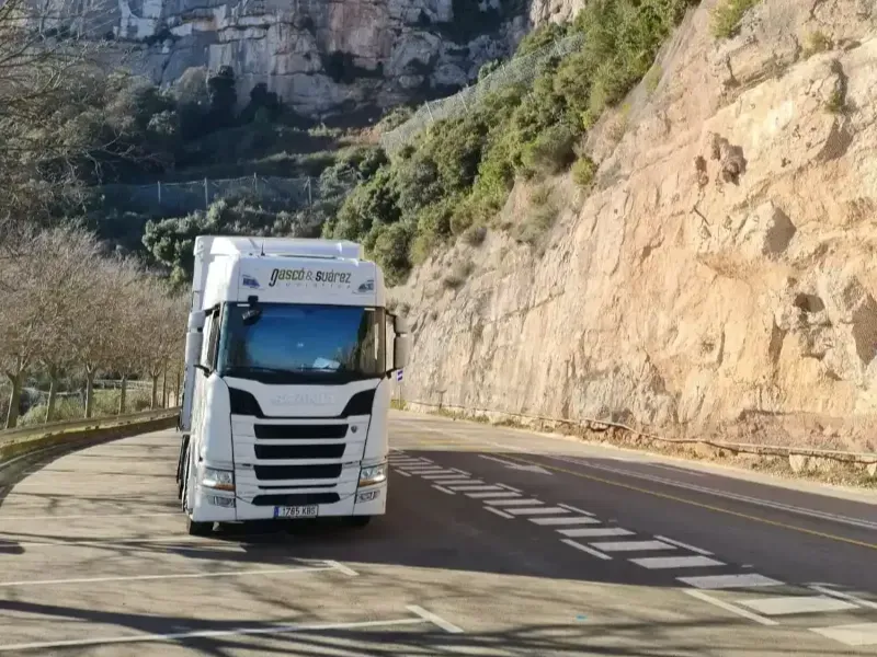 Un camión semirremolque blanco circula por una carretera sinuosa junto a una gran montaña con un fondo de cielo azul.