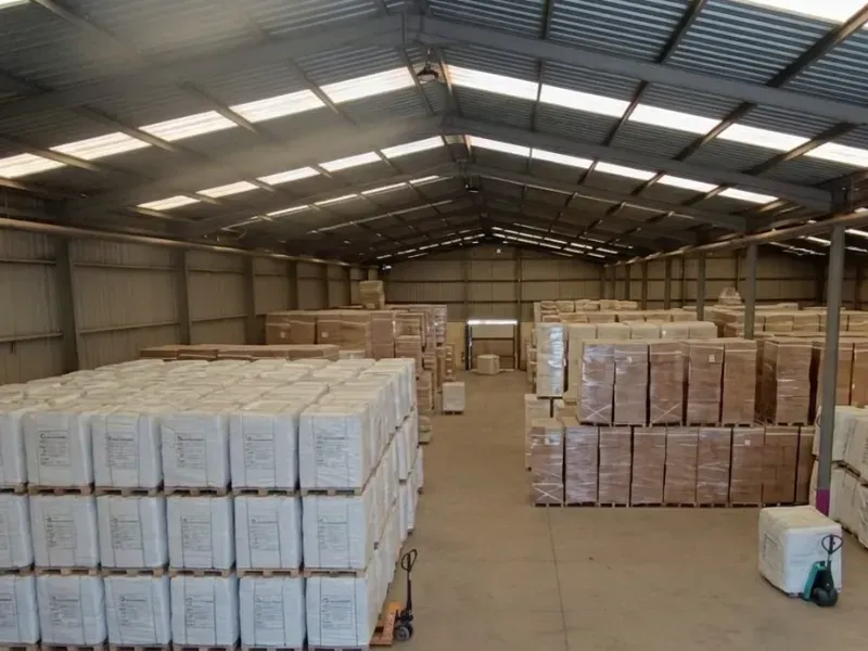 Interior del almacén con pilas de mercancía empaquetada sobre palés. Se ve una carretilla elevadora a la izquierda.