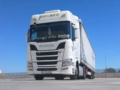 Un camión semirremolque blanco circula por una carretera sinuosa junto a una gran montaña con un fondo de cielo azul.