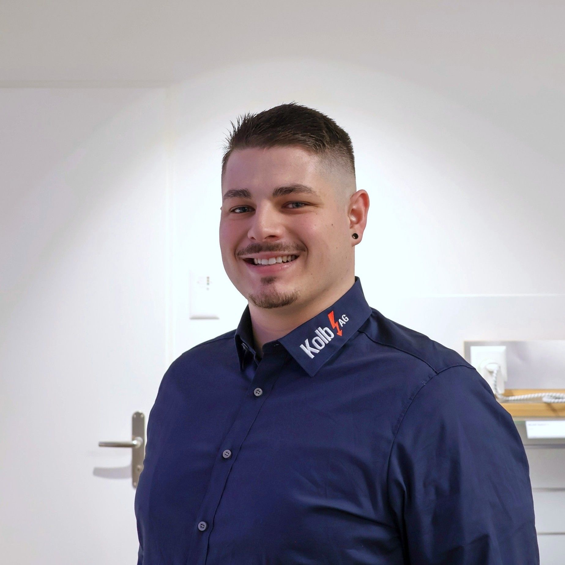 Mann mit kurzen dunklen Haaren, der ein marineblaues Hemd mit einem Logo trägt und in einem hellen Raum lächelt.