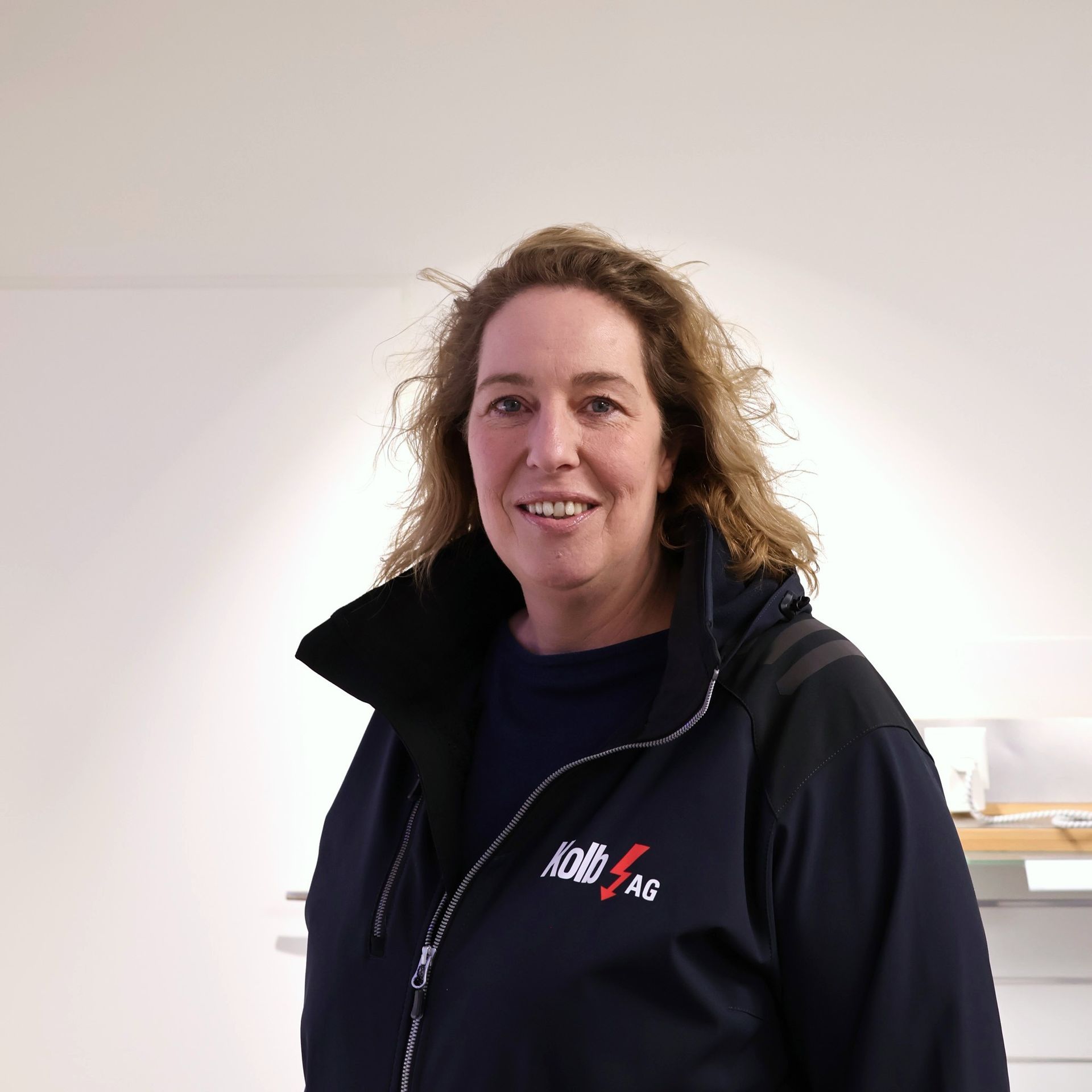 Frau mit lockigem Haar, trägt eine schwarze Jacke, lächelt und steht vor einer weißen Wand.