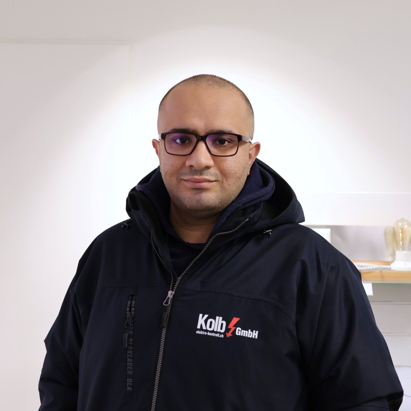 Mann mit Brille und Jacke, hält orangefarbenes Kabel und lächelt in einer Werkstatt.