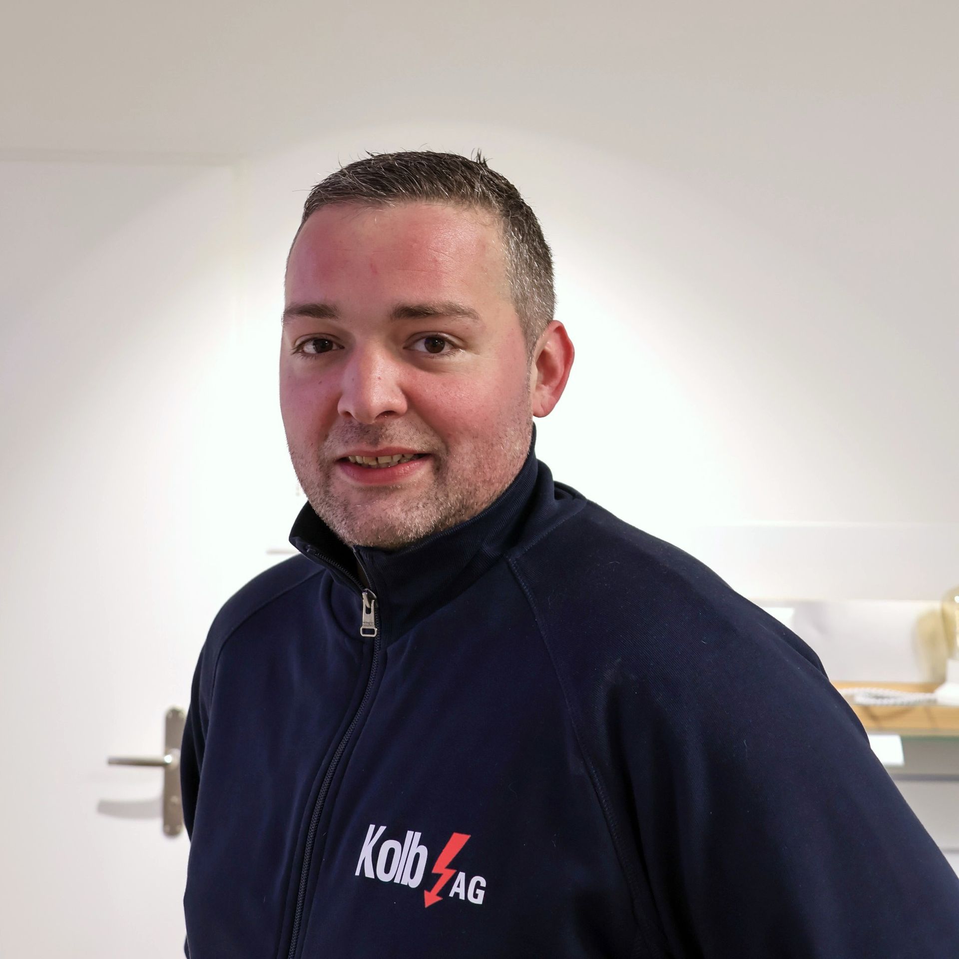 Mann in marineblauer Jacke mit „Kobag“-Logo, lächelnd in einem Innenbereich.