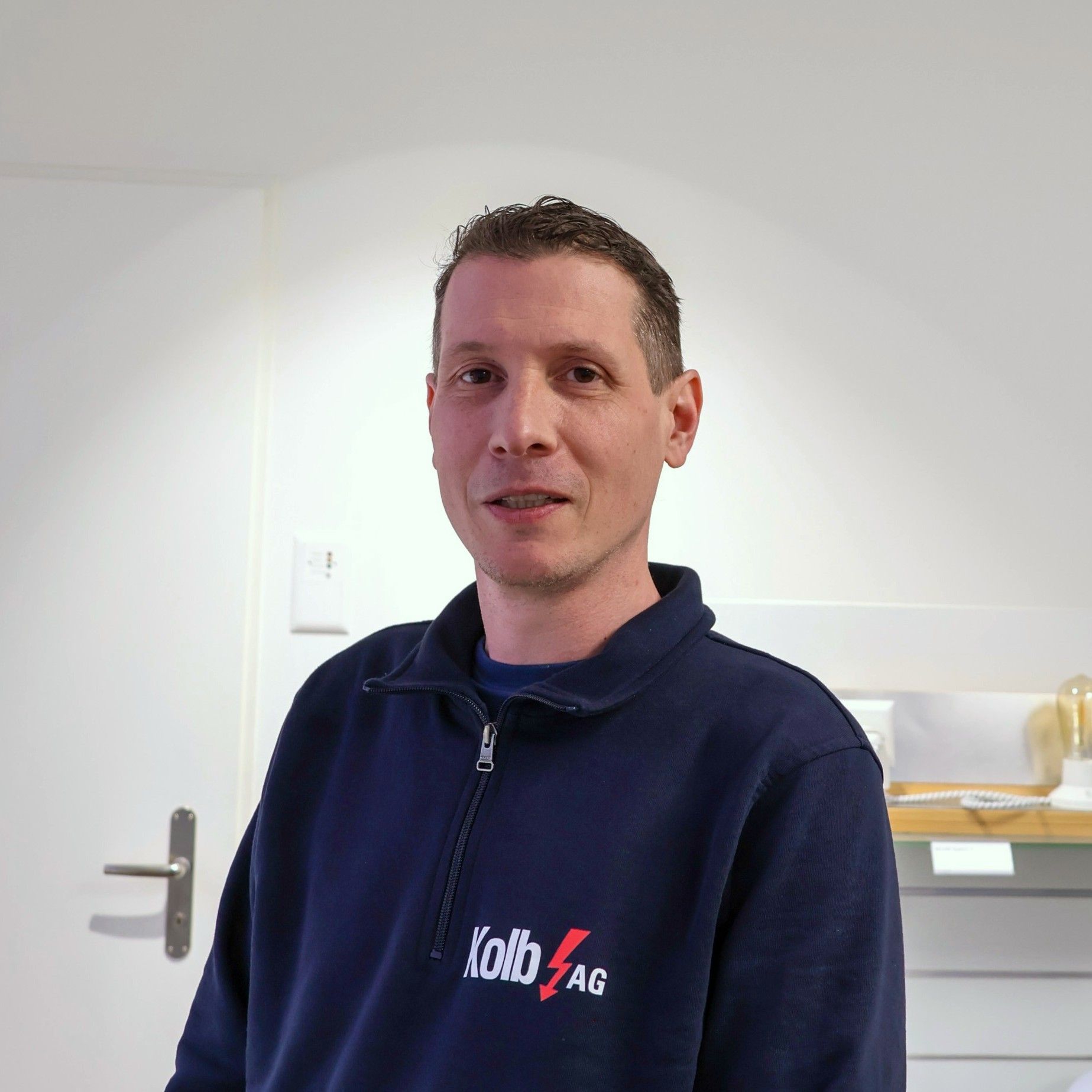 Mann im blauen Sweatshirt steht drinnen, blickt in die Kamera und lächelt leicht.