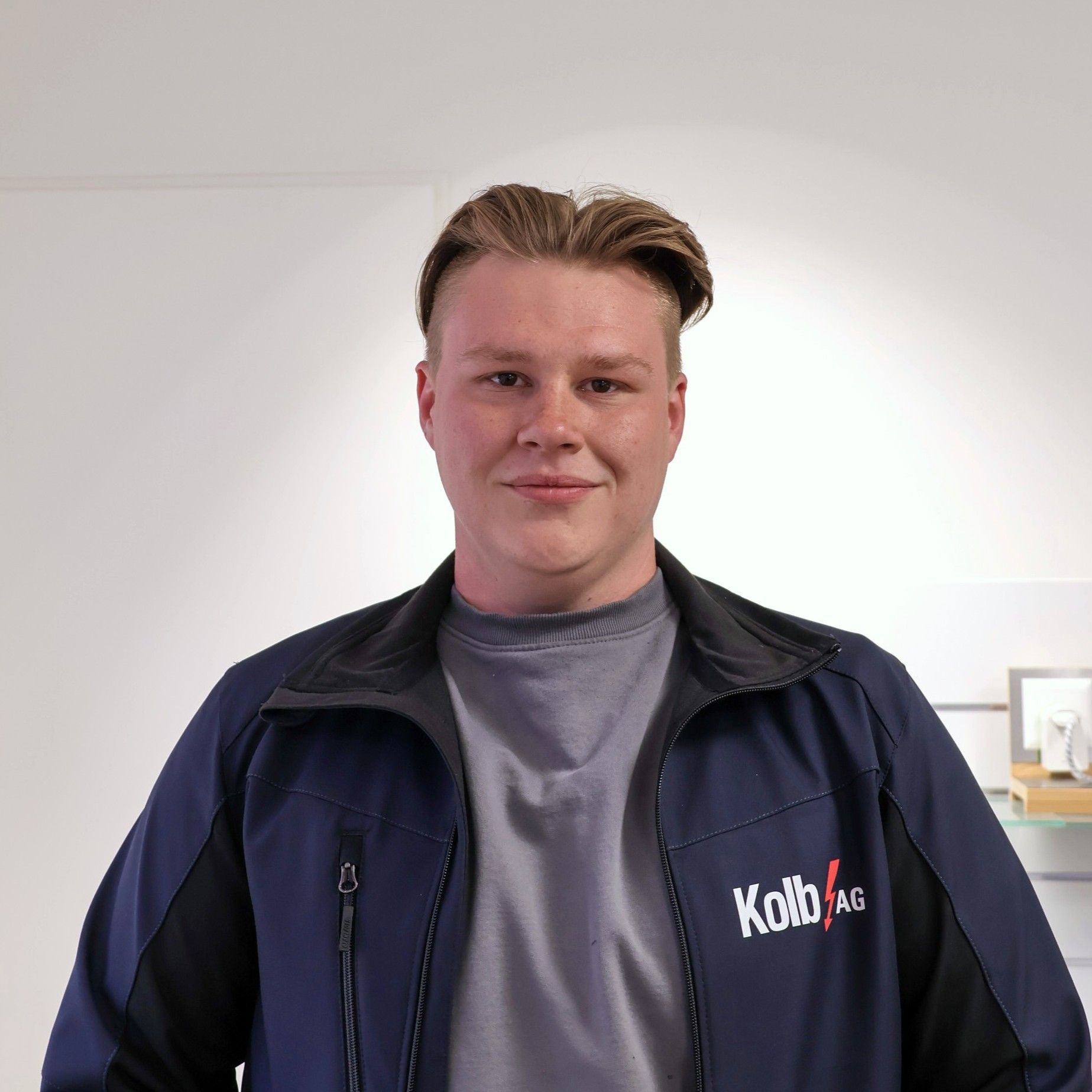 Mann in blau-schwarzer Jacke lächelt in die Kamera. Graues Hemd, helle Haut und heller Hintergrund.