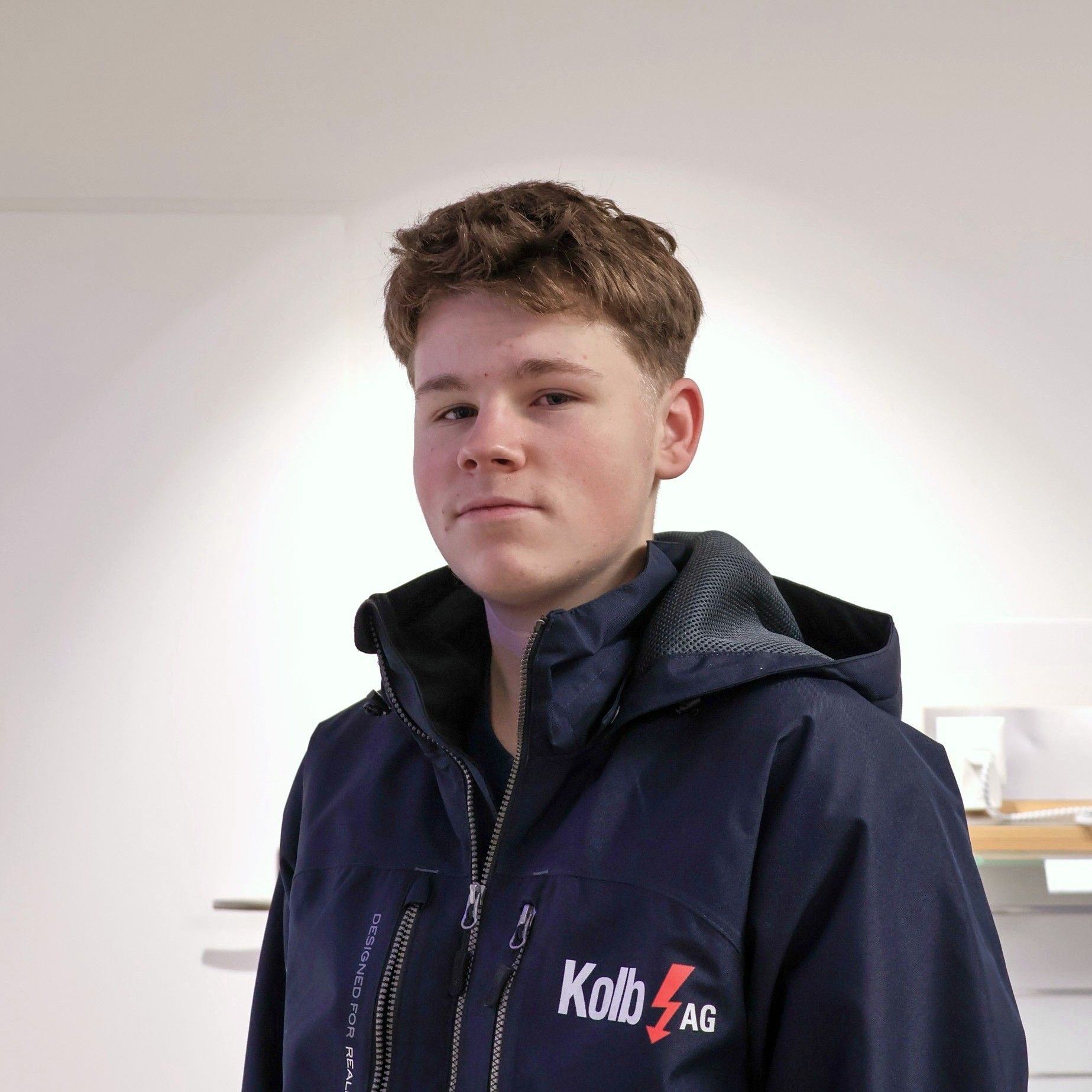 Junger Mann in einer marineblauen Jacke, braunes Haar, steht vor einer weißen Wand.