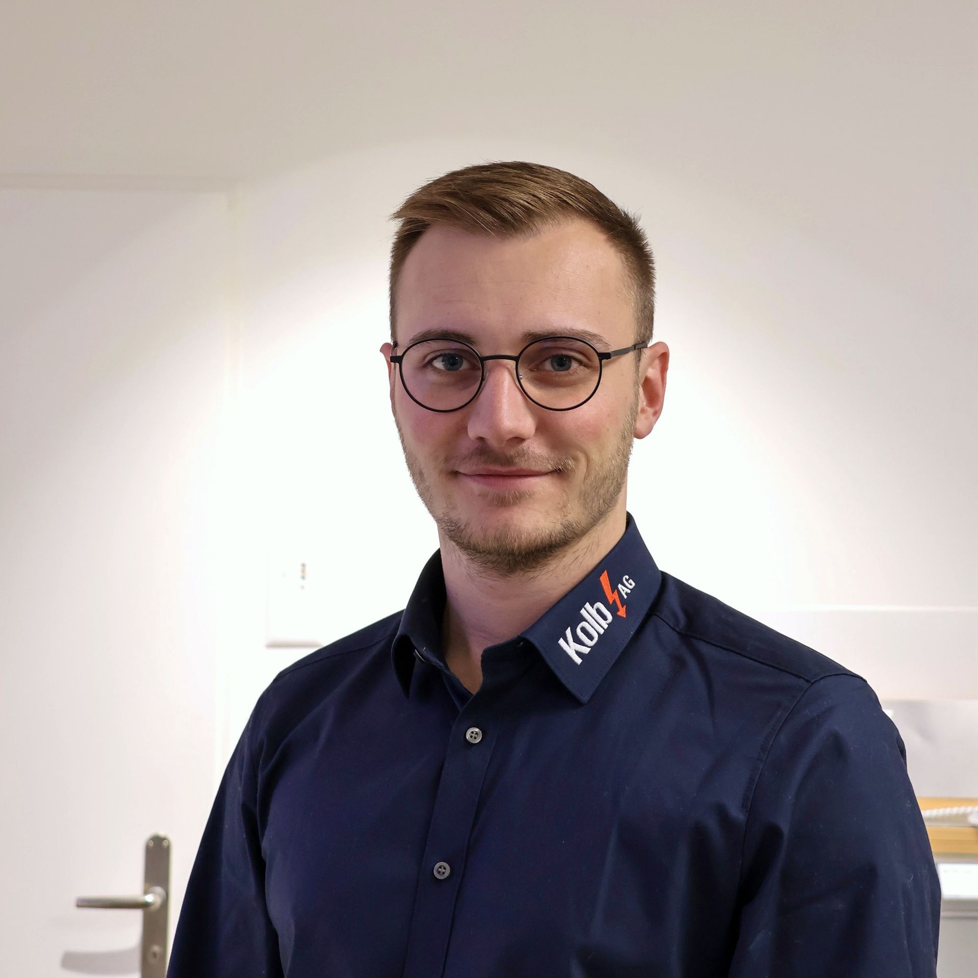 Mann mit Brille und dunkelblauem Hemd, lächelnd. Er steht in einem Büro mit weißen Wänden und trägt ein Logo auf seinem Hemd.