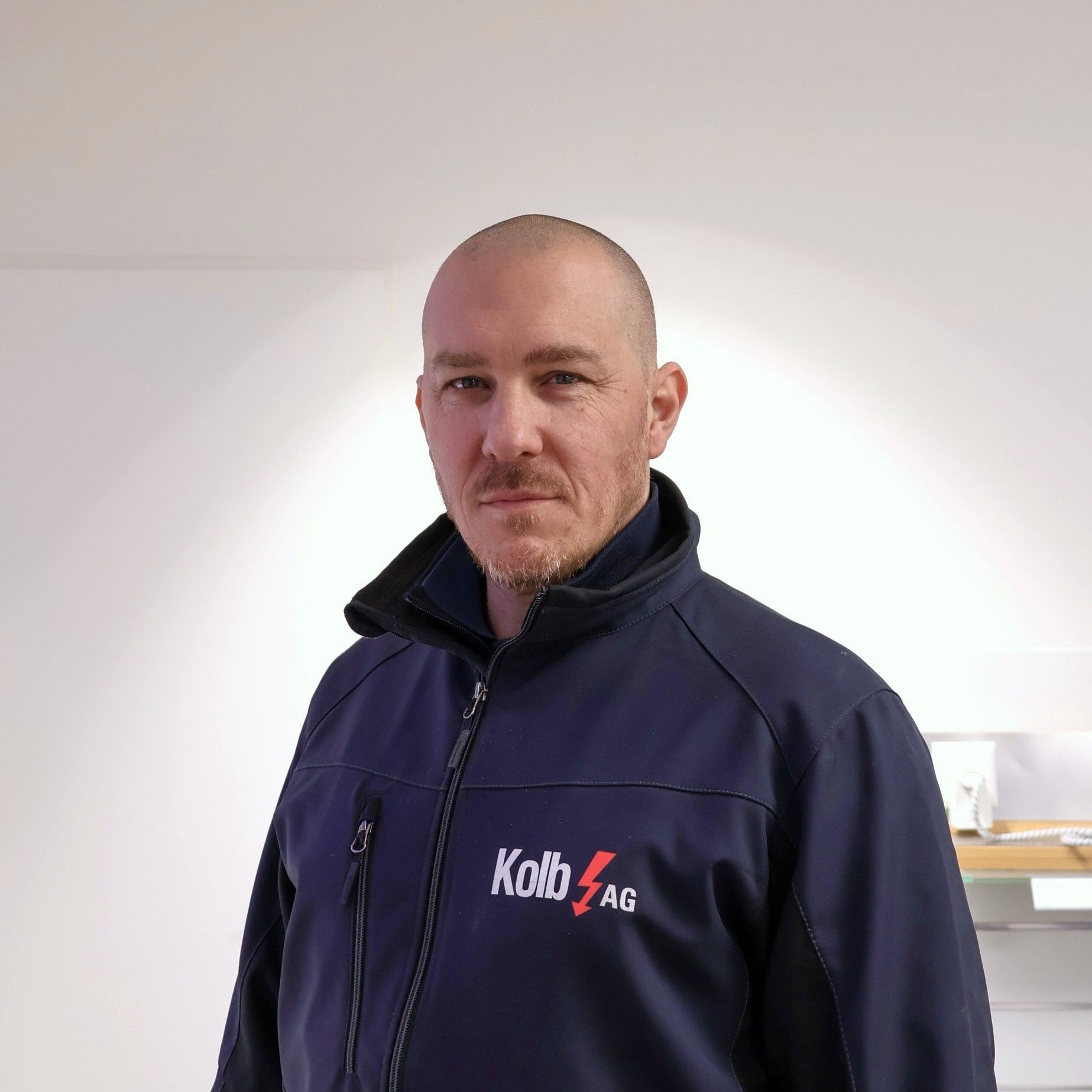 Mann mit rasiertem Kopf und marineblauer Arbeitsjacke steht in einem Raum mit weißen Wänden.