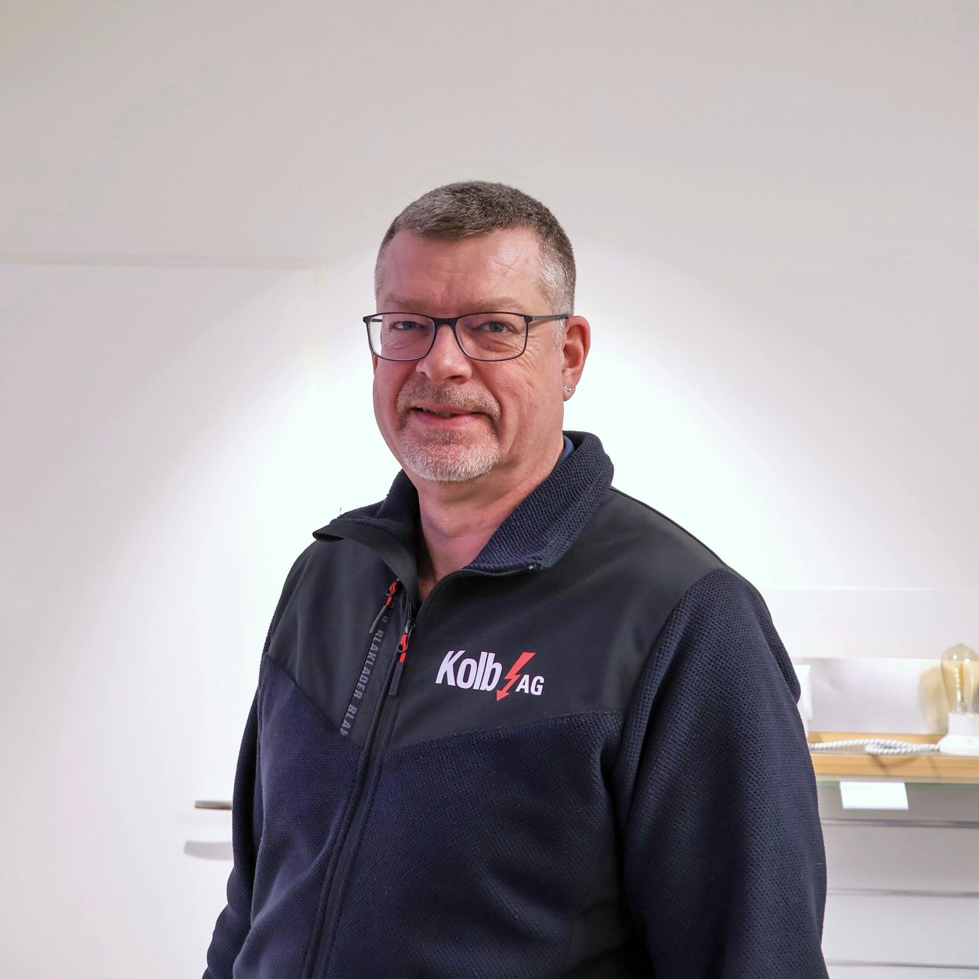 Mann mit Brille und marineblauer Jacke lächelt in einem Innenbereich.