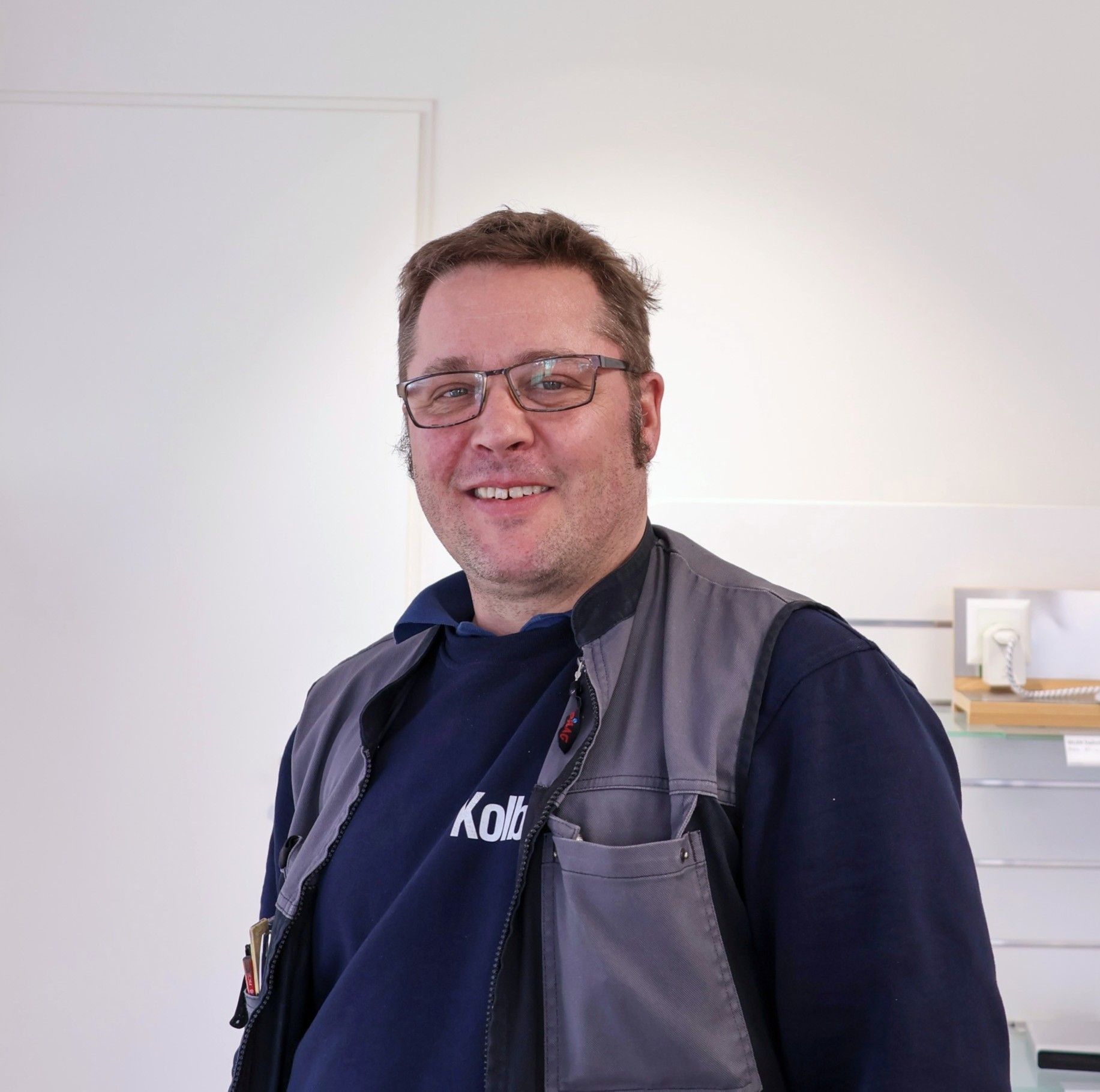 Lächelnder Mann mit Brille, der eine graue Weste über einem blauen Sweatshirt trägt und in einem Gebäude neben einer weißen Wand steht.