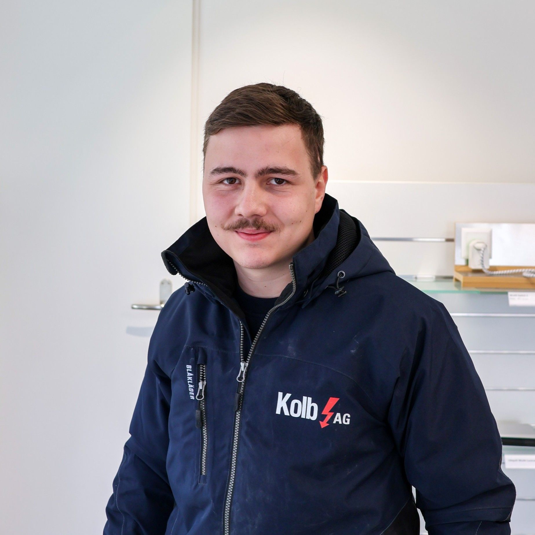 Mann in blauer Jacke mit Logo steht mit neutralem Gesichtsausdruck im Haus.