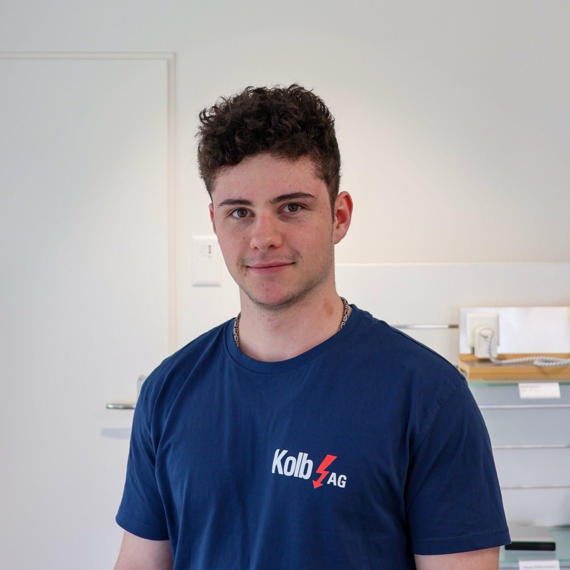 Ein junger Mann in einem blauen T-Shirt steht lächelnd in einem Raum mit weißen Wänden. Auf dem T-Shirt ist ein Logo abgebildet und er hat lockiges Haar.