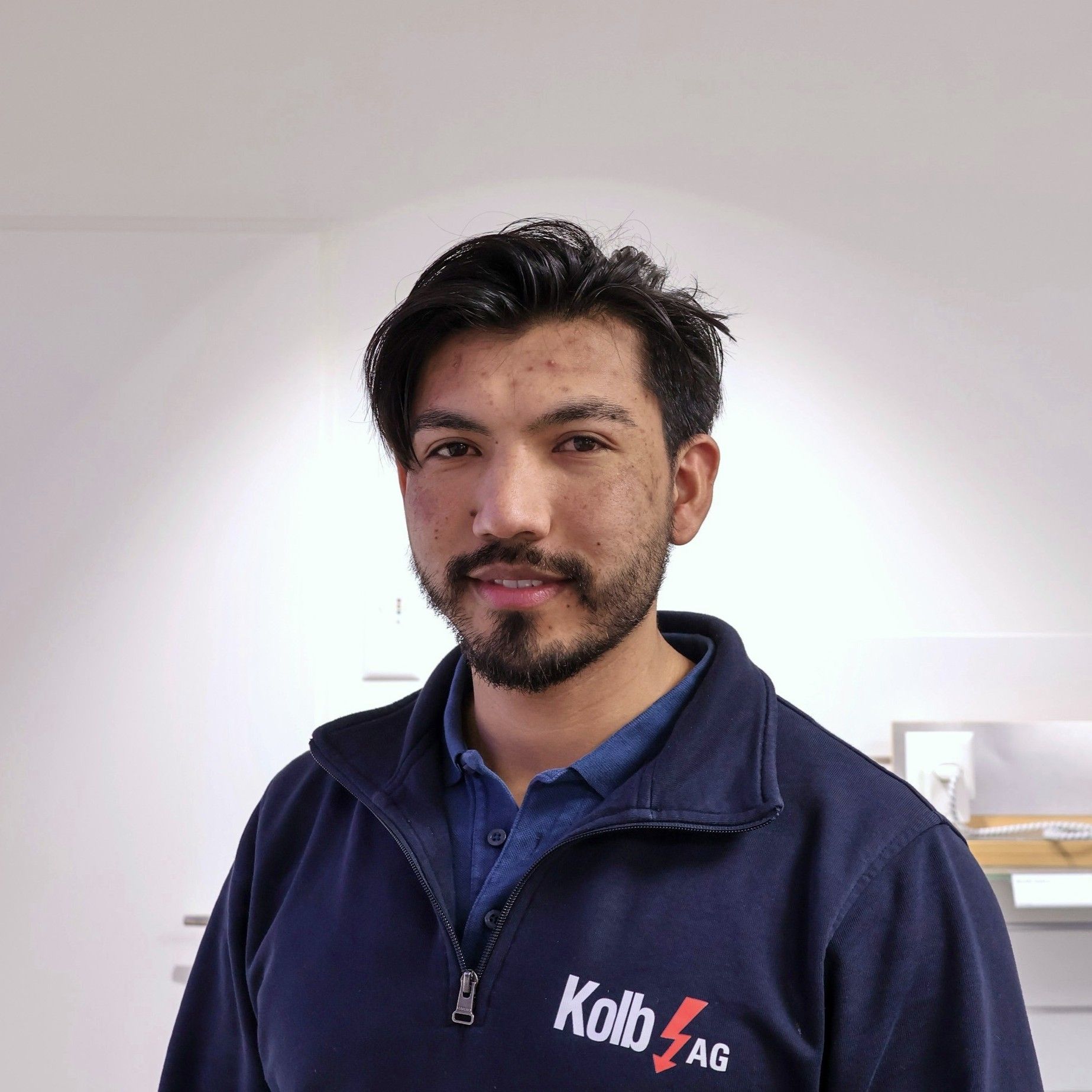 Mann mit dunklem Haar und Bart in einer blauen Jacke der Kolb AG, im Innenbereich.