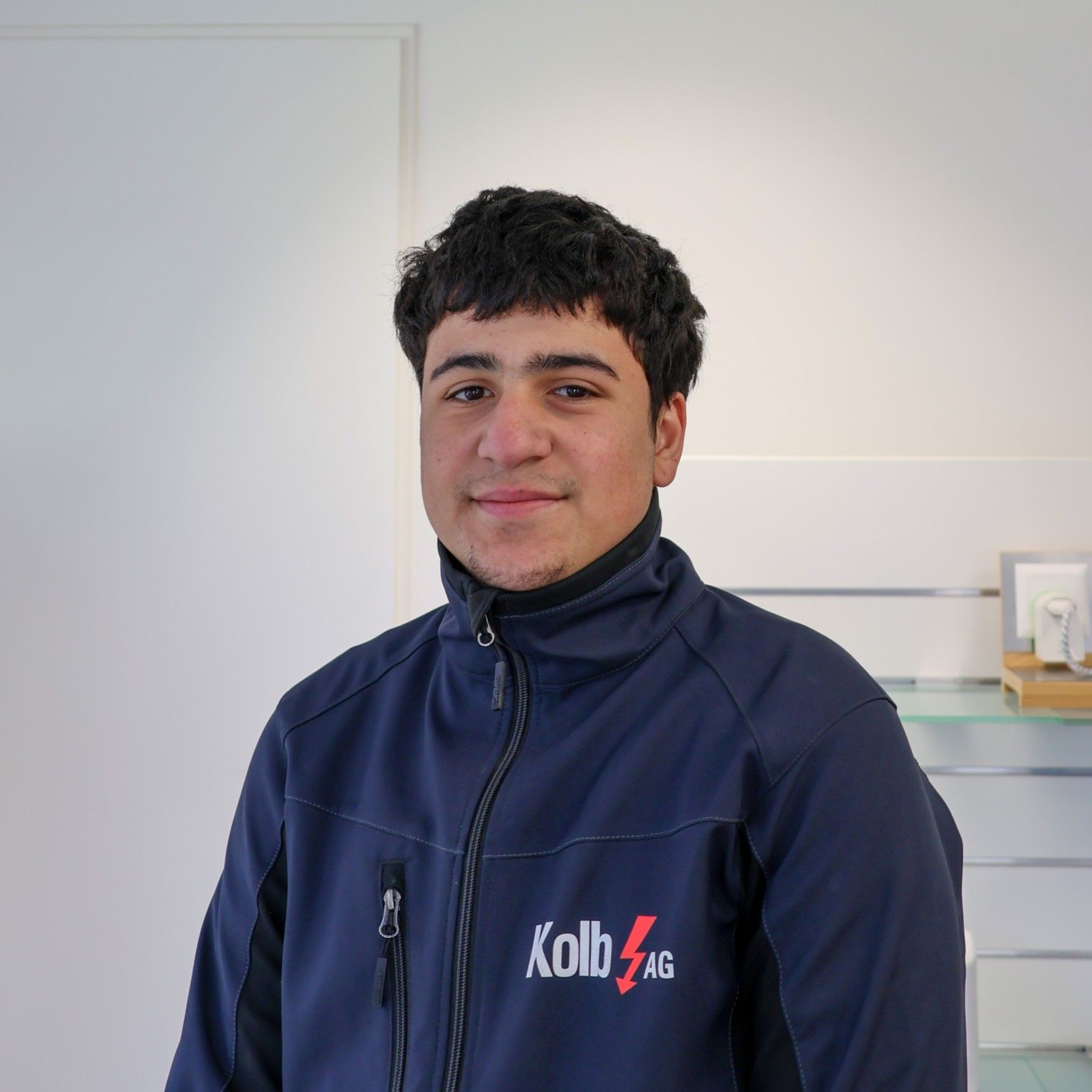 Junger Mann mit lockigem braunem Haar in einer blauen Jacke, lächelnd, steht drinnen.