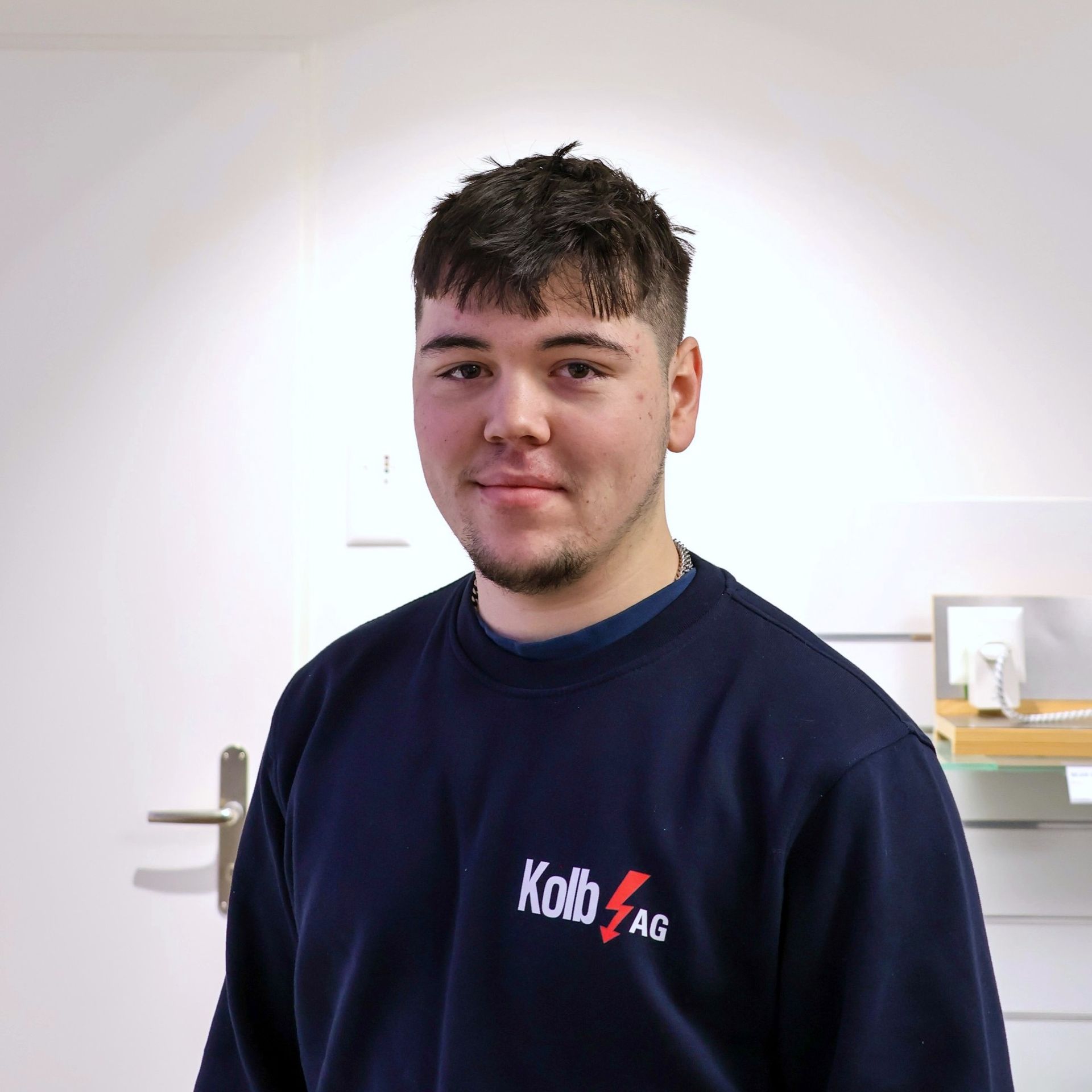 Junger Mann im blauen Sweatshirt, lächelnd, steht in einem Büro, Logo auf seinem Hemd.