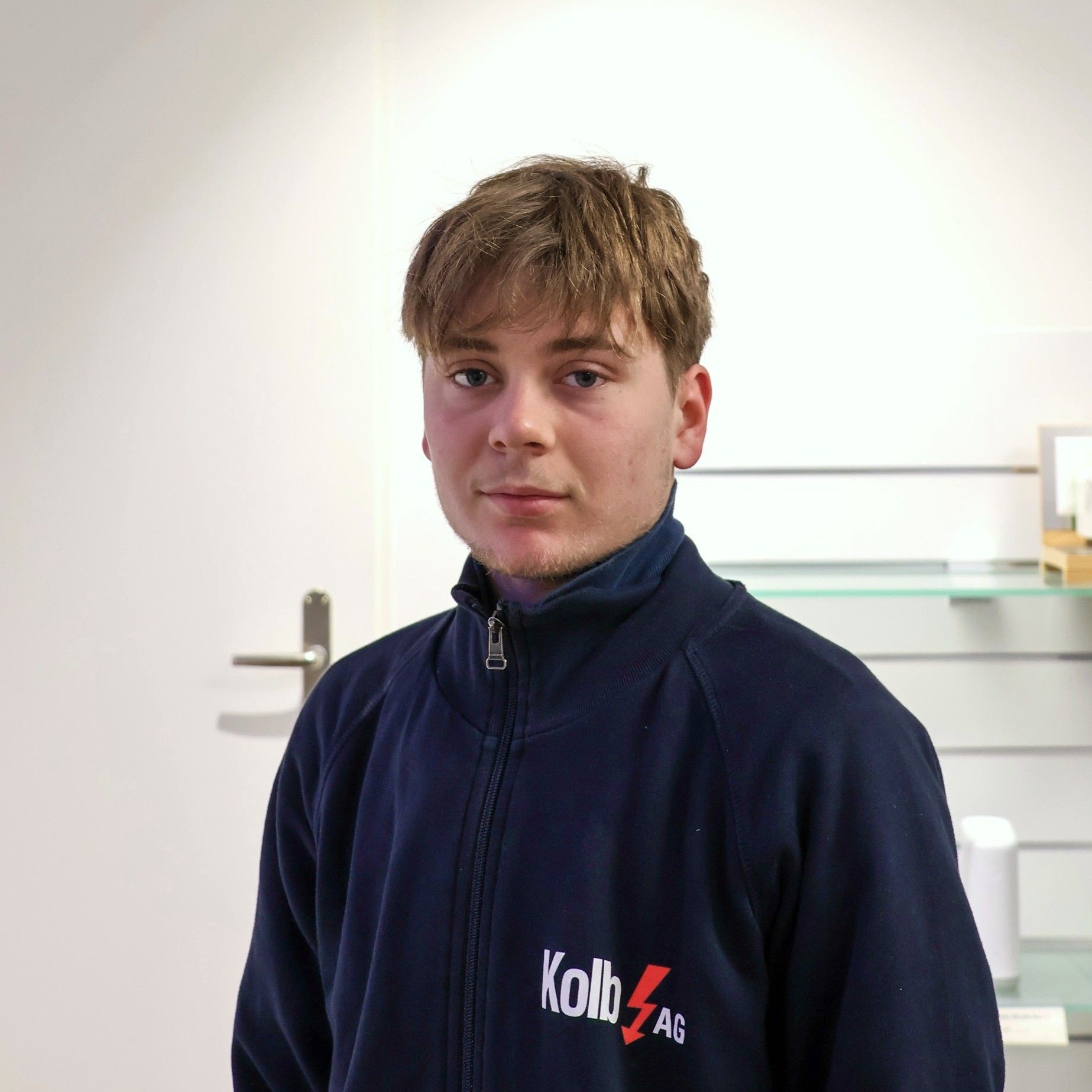 Junger Mann in blauem Sweatshirt mit Logo, drinnen stehend.