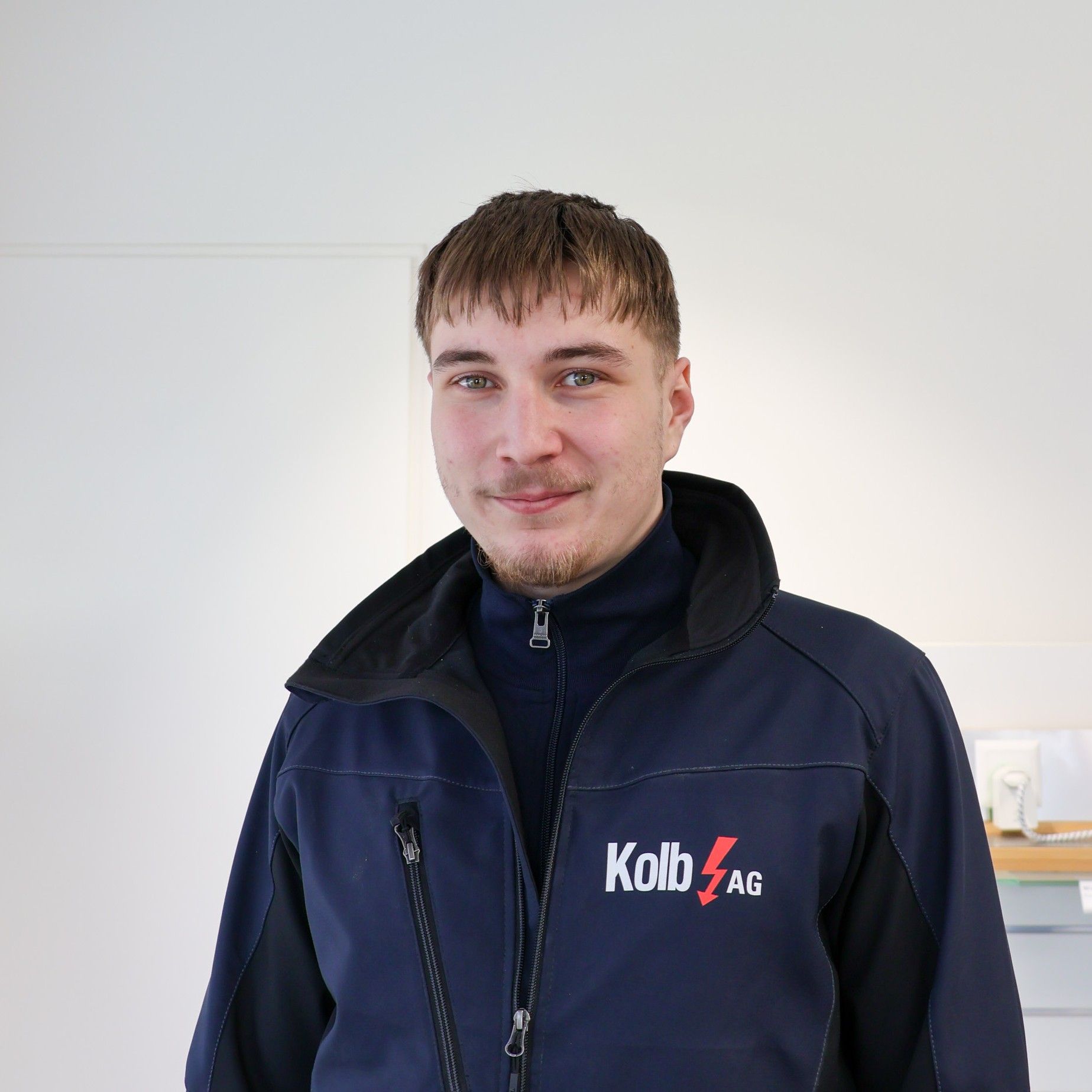 Mann in blauer Jacke mit „Kolb AG“-Logo, lächelnd vor einer weißen Wand.
