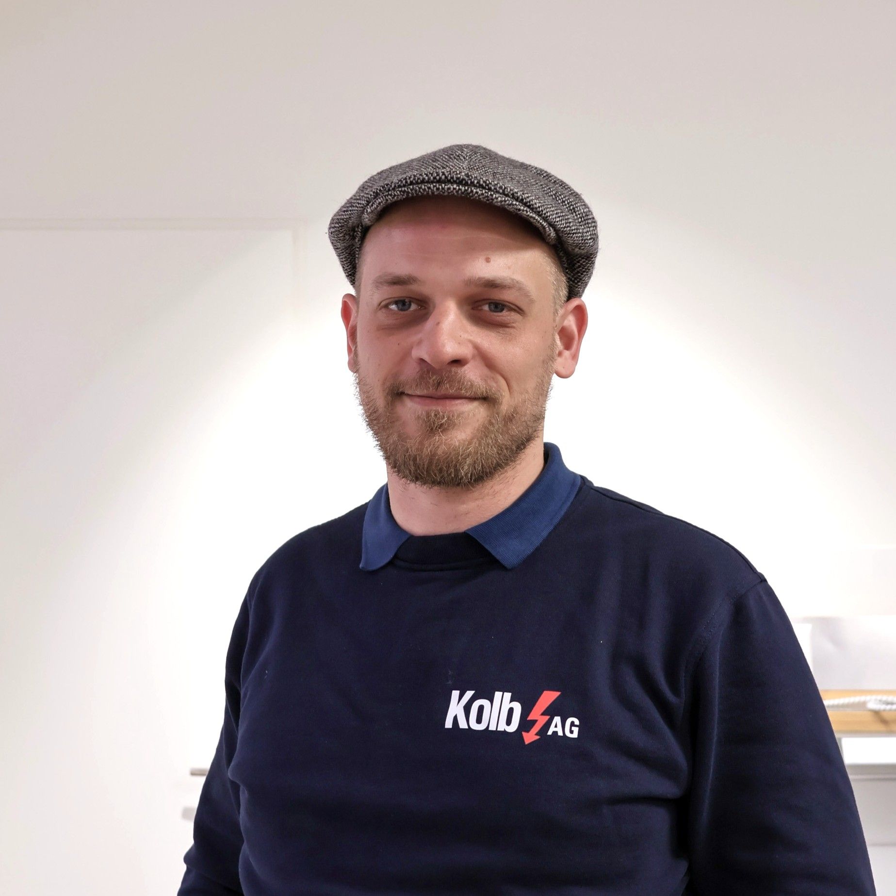 Mann mit Mütze und blauem Sweatshirt mit „Kolb“-Logo, lächelnd in einem Raum.