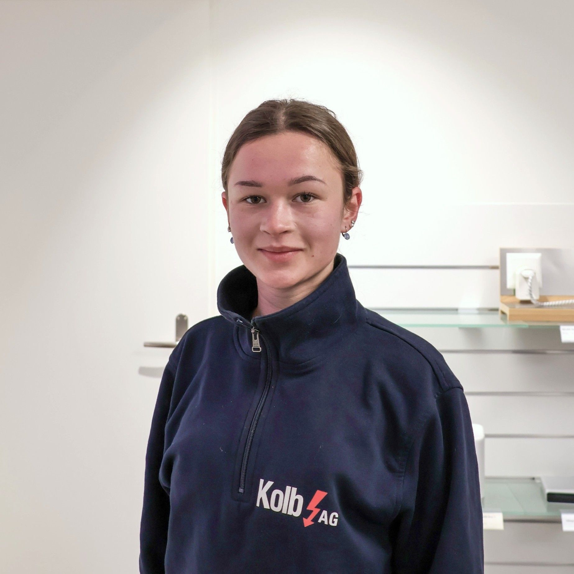 Frau in blauem Sweatshirt mit Logo, steht vor einer weißen Wand und Regalen.