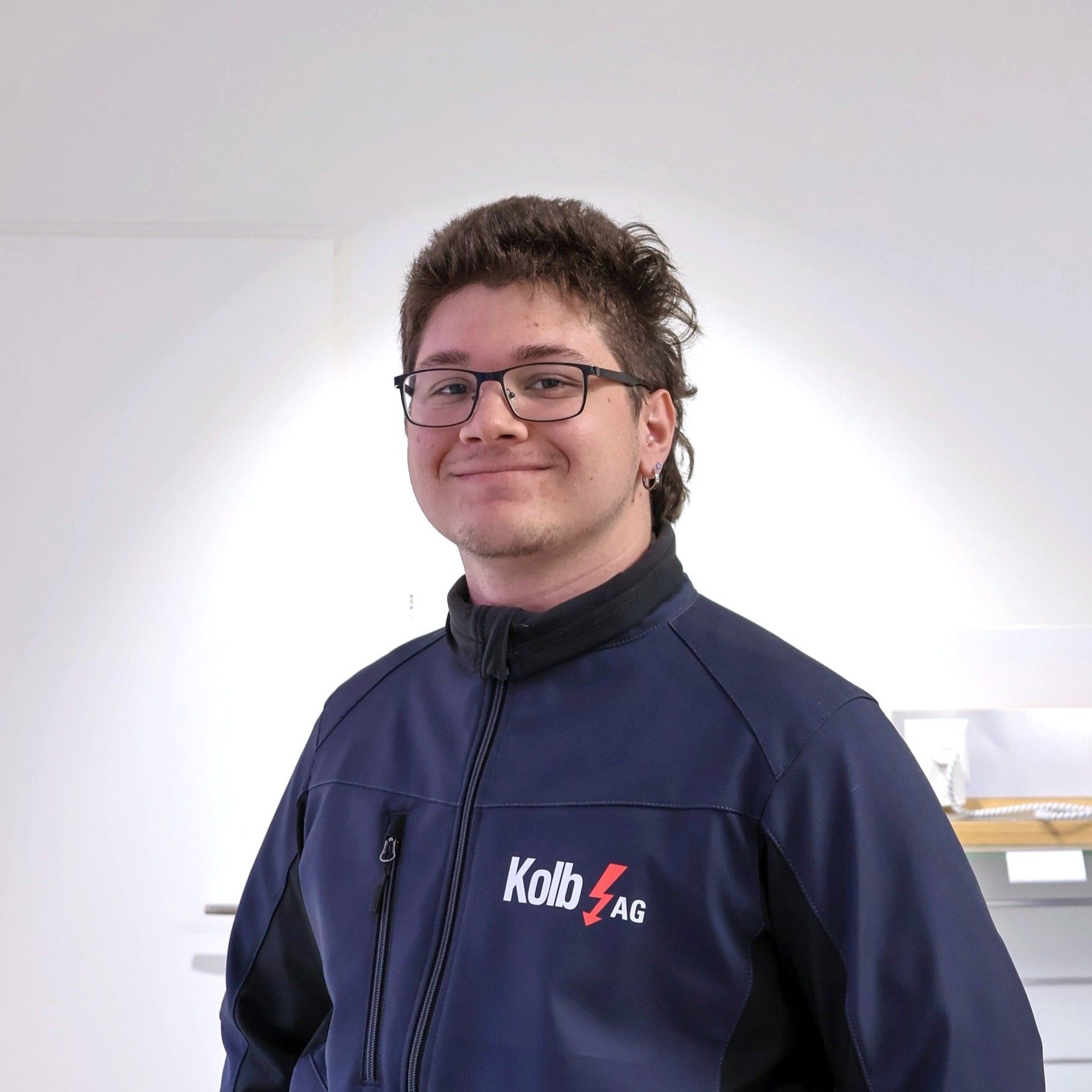 Mann in blauer Arbeitsjacke mit Brille lächelt in einem weißen Raum.