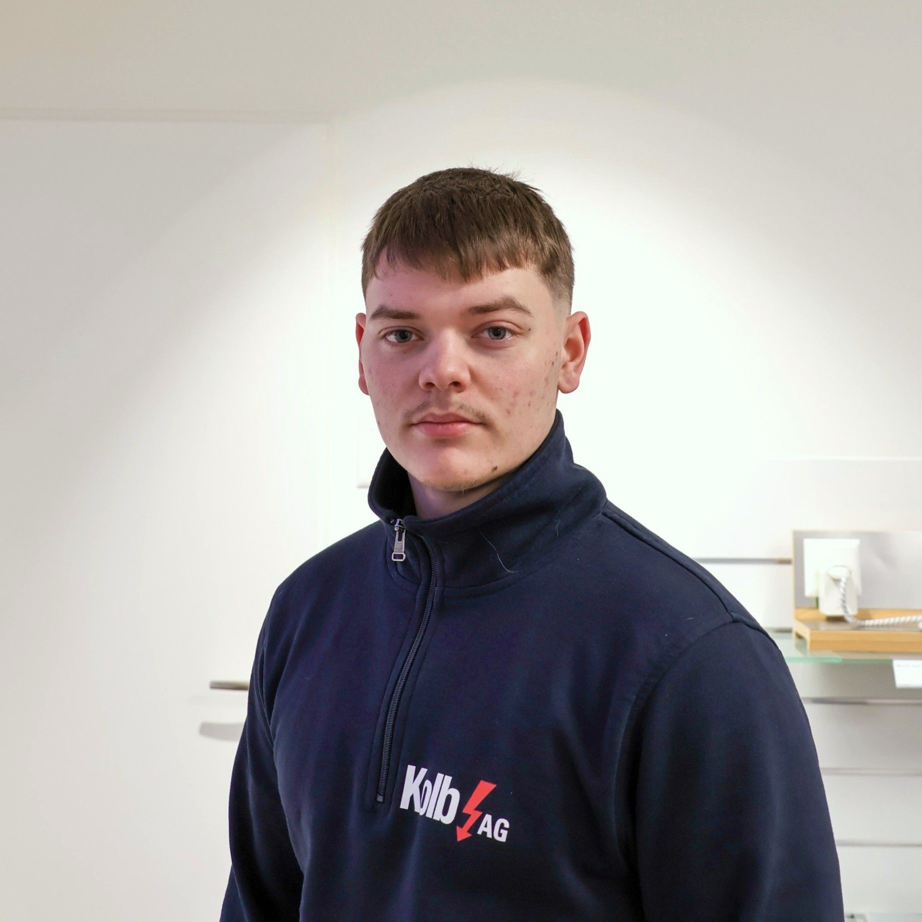 Junger Mann in blauem Reißverschluss, KB4 AG-Logo, steht drinnen, neutraler Gesichtsausdruck.