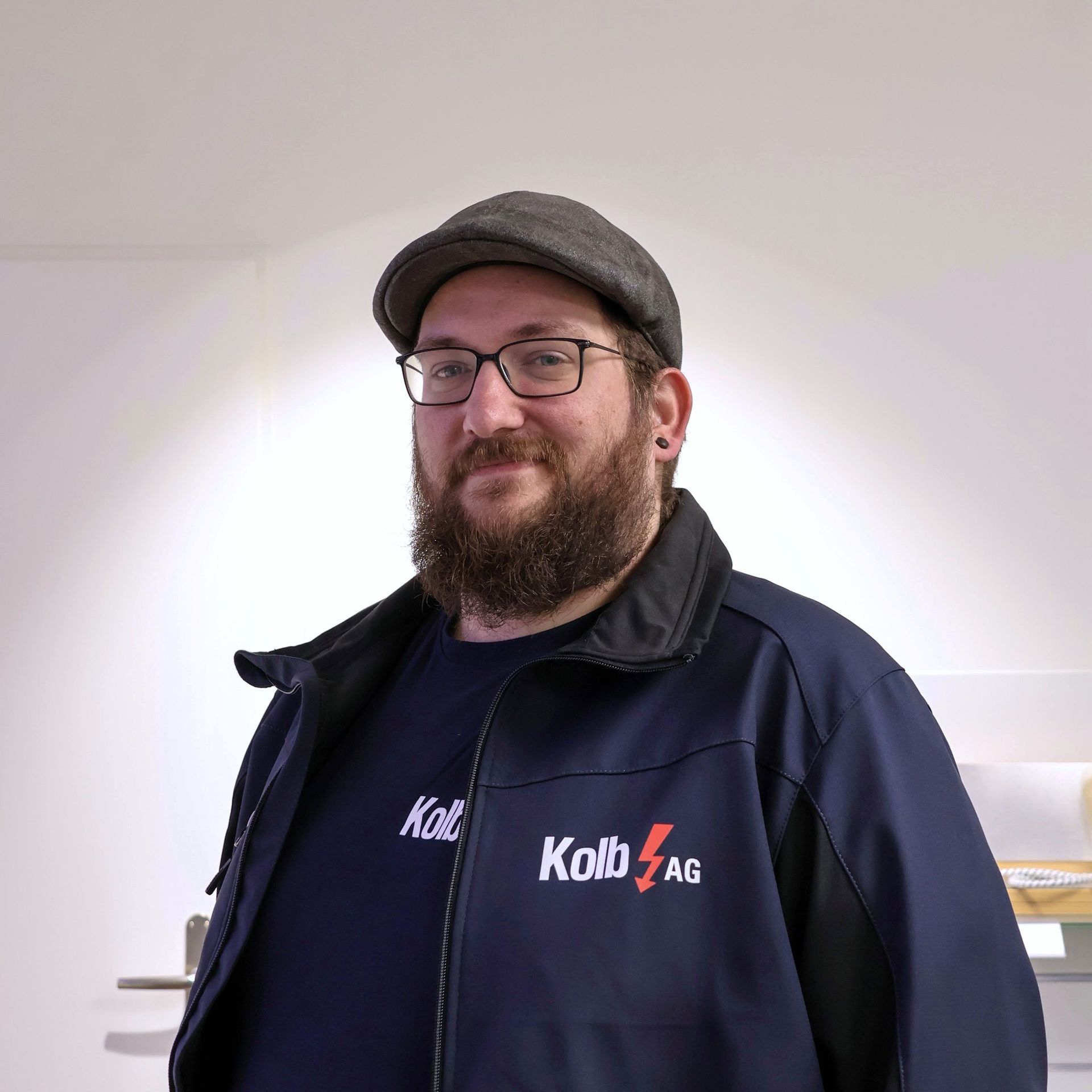 Mann mit Bart und Brille, der eine Jacke und eine Mütze von Kolb AG trägt und in einem Raum mit weißen Wänden lächelt.