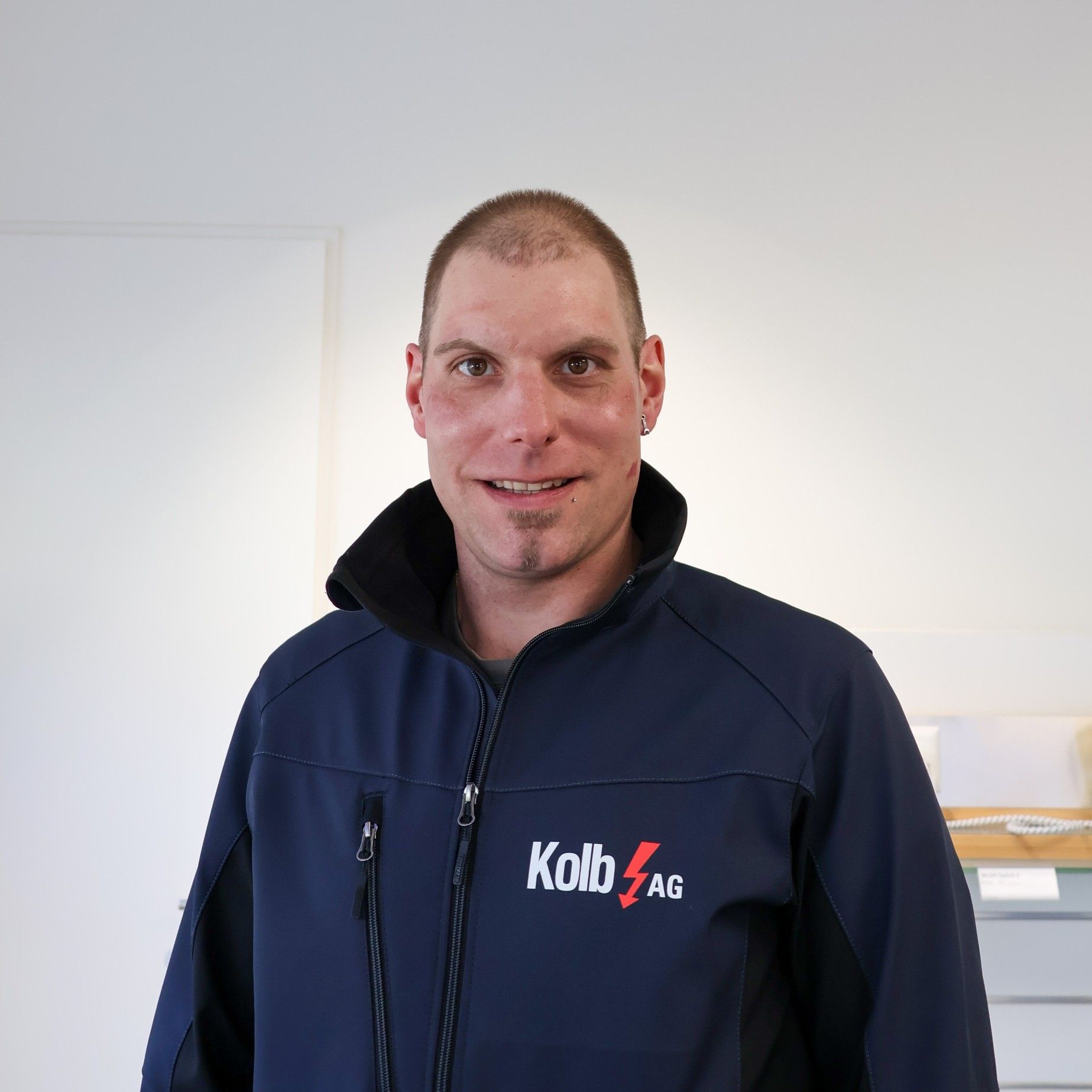 Mann in blauer Kolb AG-Jacke lächelt, steht drinnen.