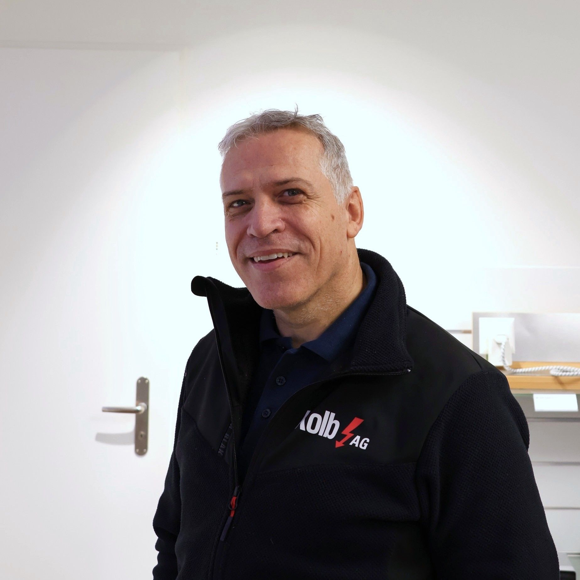 Mann in schwarzer Jacke lächelt in die Kamera. Steht in einem Raum mit weißen Wänden.