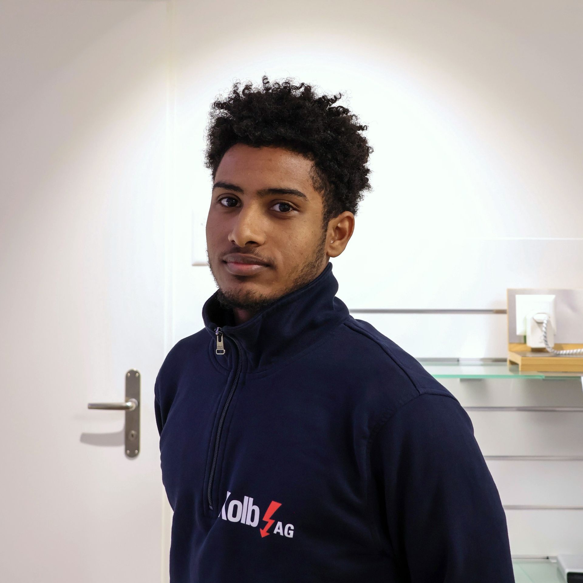 Mann mit lockigem Haar, trägt ein blaues Sweatshirt mit Reißverschluss und blickt in die Kamera.