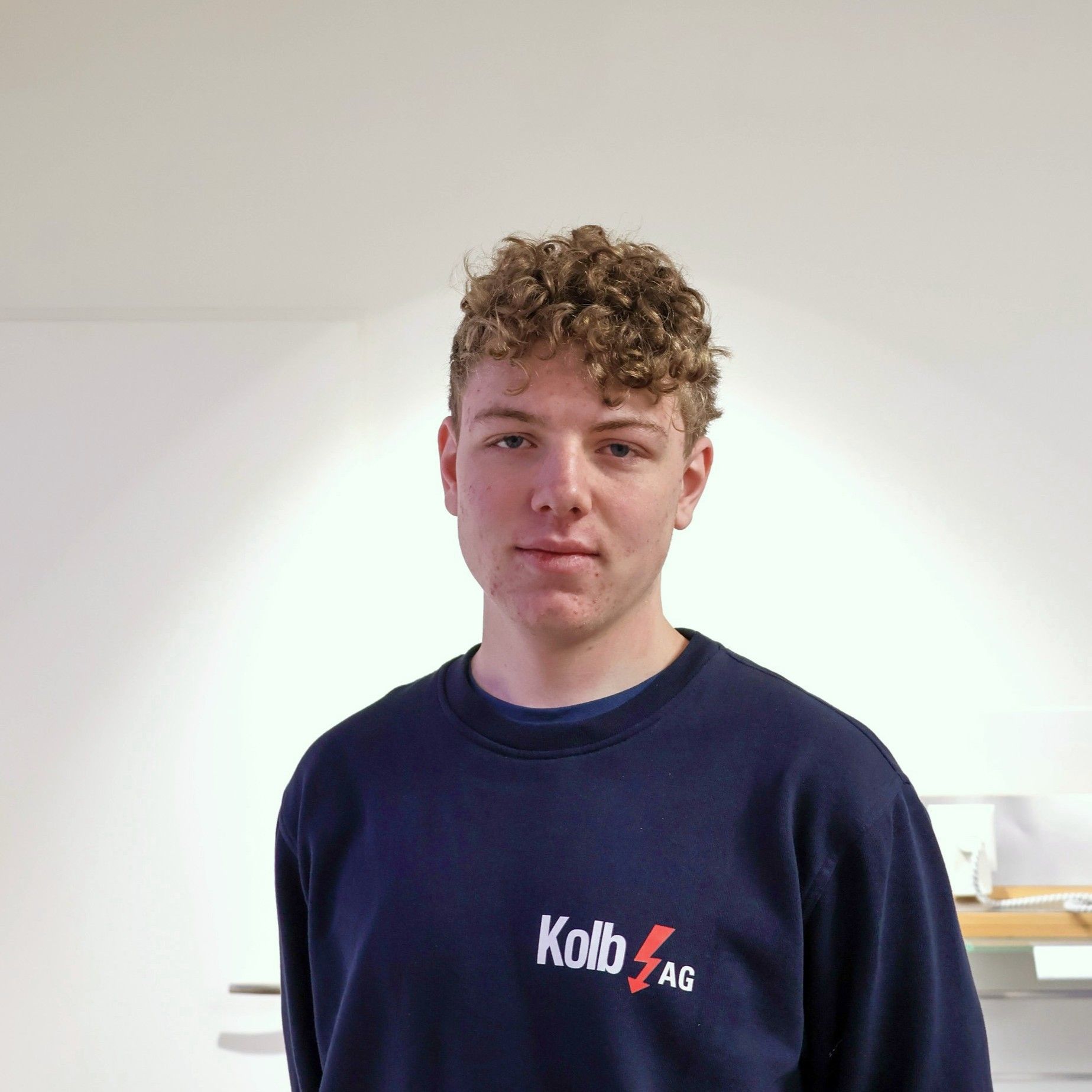 Junger Mann mit lockigem Haar, der einen marineblauen Pullover mit einem Firmenlogo trägt und im Haus steht.
