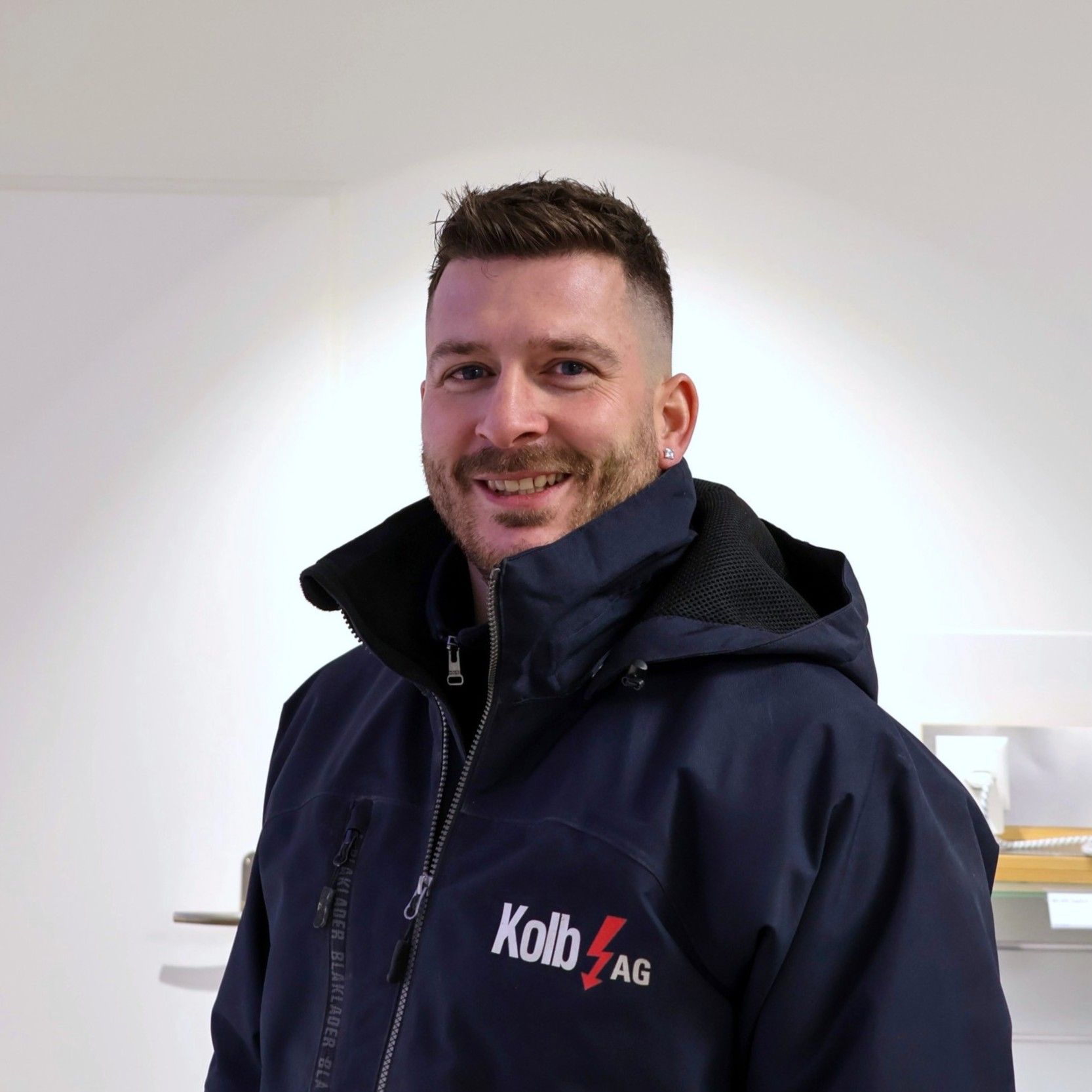 Mann mit kurzen braunen Haaren, trägt eine dunkelblaue Jacke mit dem Logo der „Kolb AG“. Lächelnd.