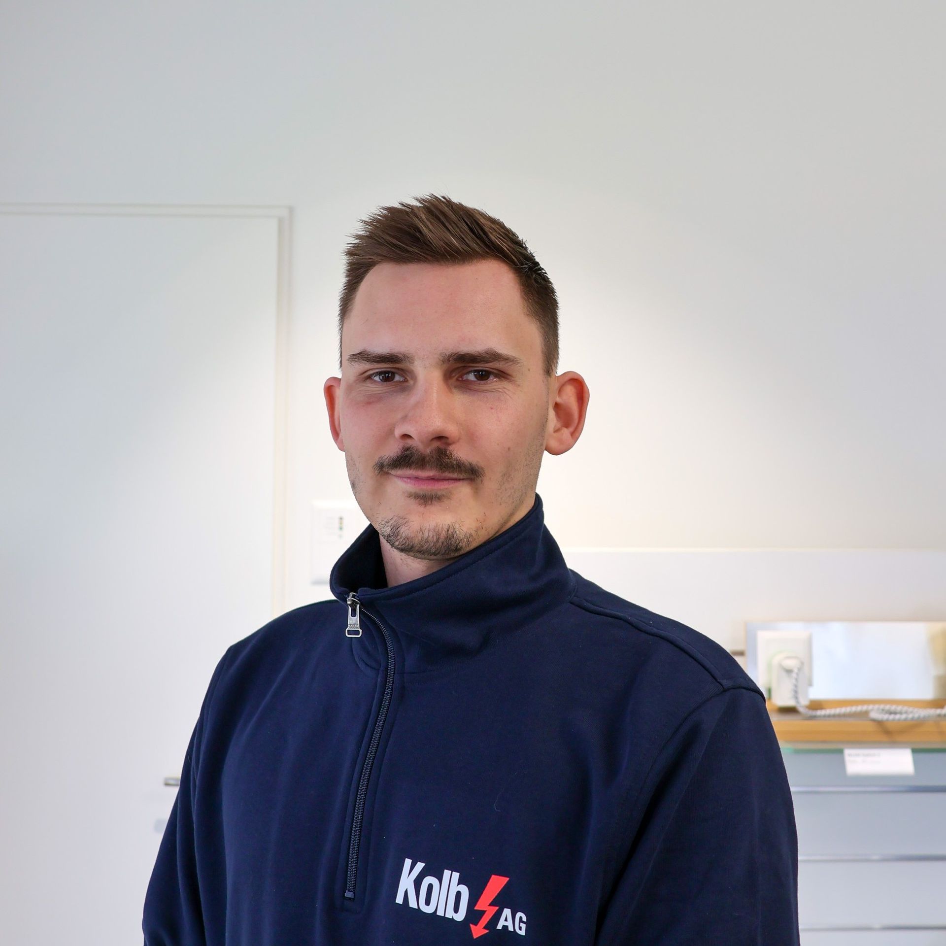 Mann mit Schnurrbart, blaue Jacke mit „Kolb AG“-Logo, in einem weißen Raum.