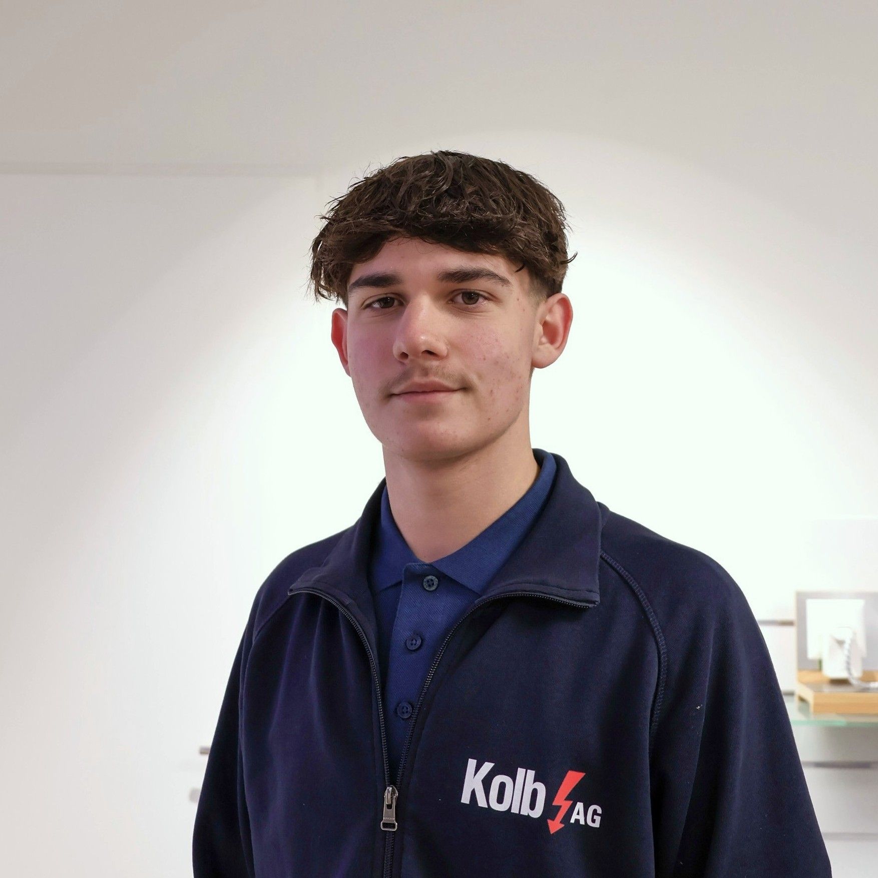 Junger Mann in einer blauen Jacke mit dem Logo der Kolb AG, lächelnd in einem Innenbereich.