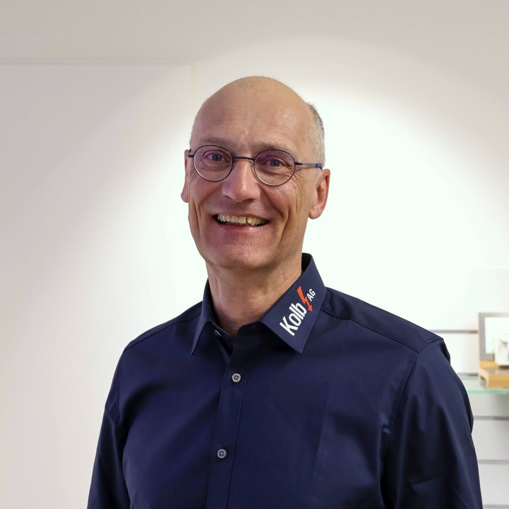 Ein lächelnder, kahlköpfiger Mann in einem blauen Hemd und mit Brille steht drinnen.