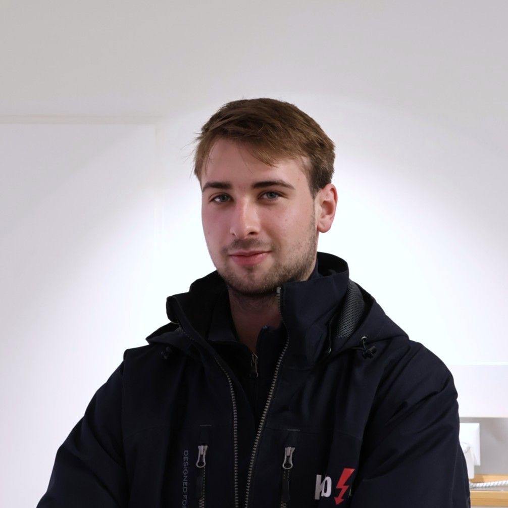 Junger Mann lächelt im Freien, trägt ein weißes Hemd mit einem „Kolb“-Logo; im Hintergrund ein weißer Lieferwagen und ein Gebäude.