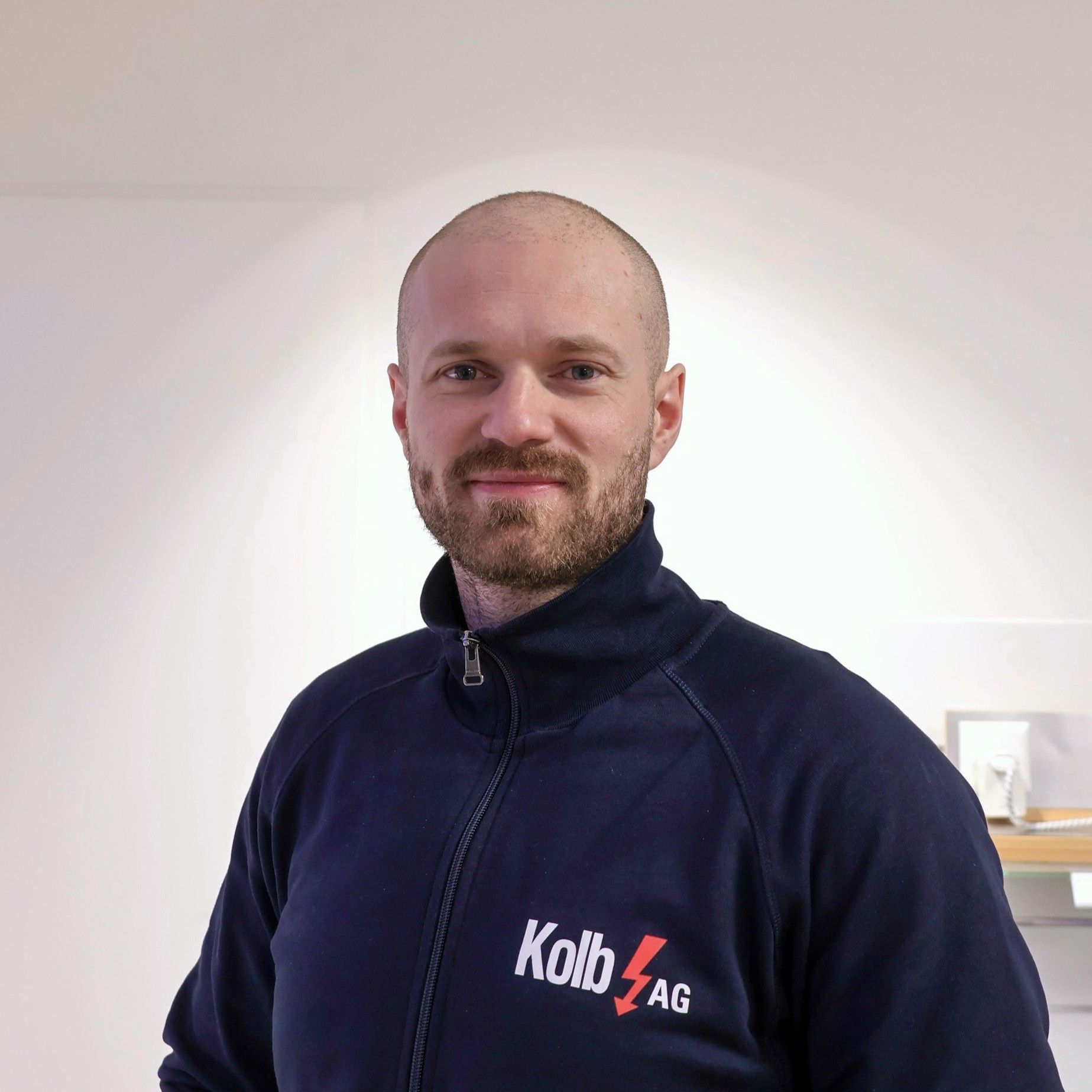 Ein lächelnder Mann in einer blauen Reißverschlussjacke mit einem Logo. Innenaufnahme, neutraler Hintergrund.