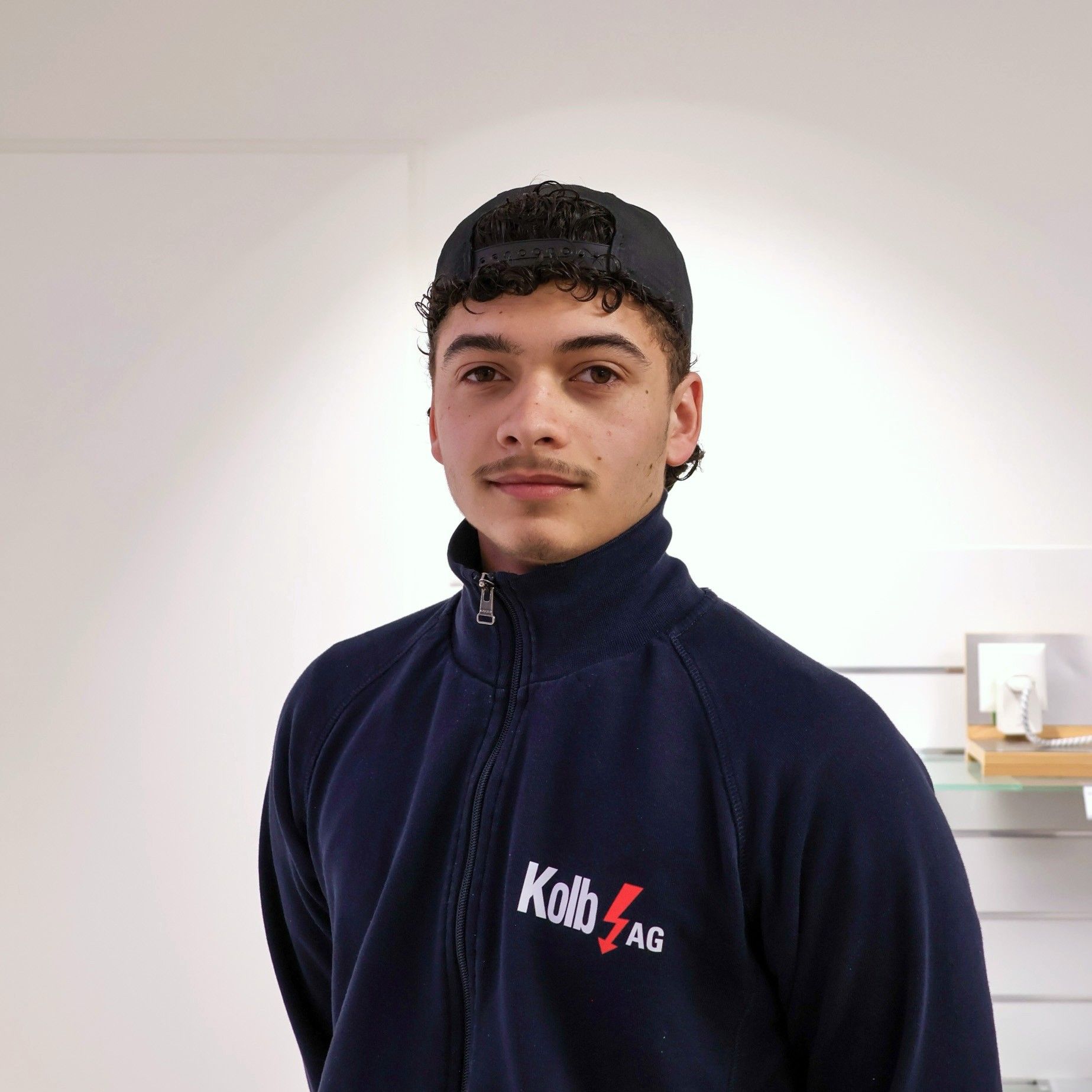 Junger Mann in dunkelblauer Jacke und schwarzer Mütze, lächelnd. Stehend in einem Raum mit weißen Wänden.