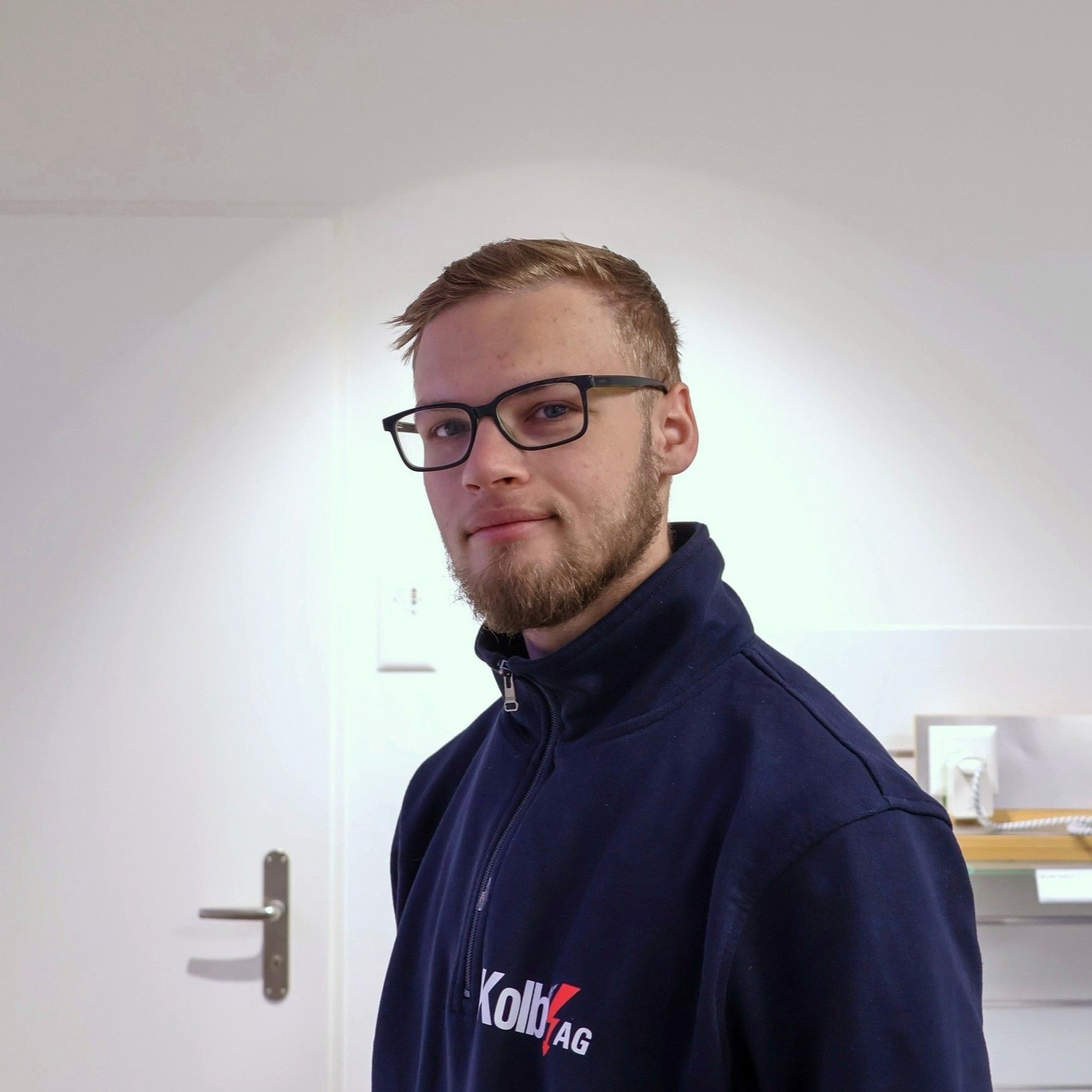 Mann mit Brille und Bart, der eine blaue Arbeitsjacke trägt, lächelt und in einem weißen Raum steht.