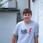 Junge Person in grauem T-Shirt, lächelnd, an eine weiße Wand gelehnt.