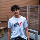 Ein junger Mann mit lockigem Haar in einem hellgrauen T-Shirt lächelt vor einem hölzernen Hintergrund.