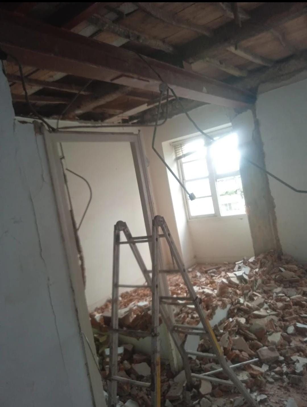 Habitación dañada, con el techo derrumbado, escombros en el suelo, una escalera y luz intensa entrando por la ventana