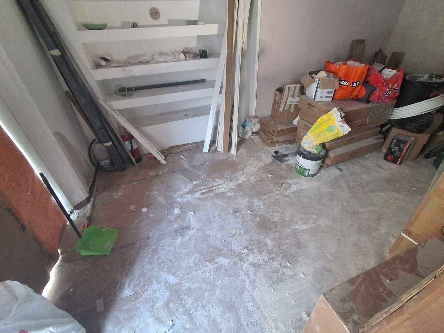 Cuarto de servicio desordenado, con escaleras, artículos de limpieza y bolsas de suministros sobre el suelo polvoriento.