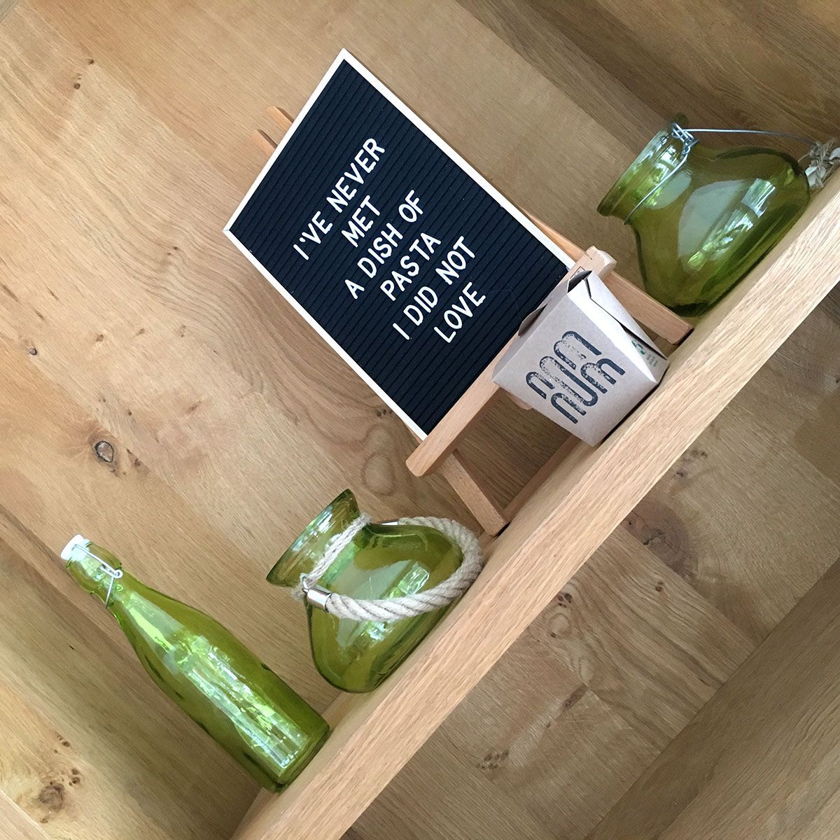 Houten plank met groene glazen flessen en potten, en een bordje met de tekst: