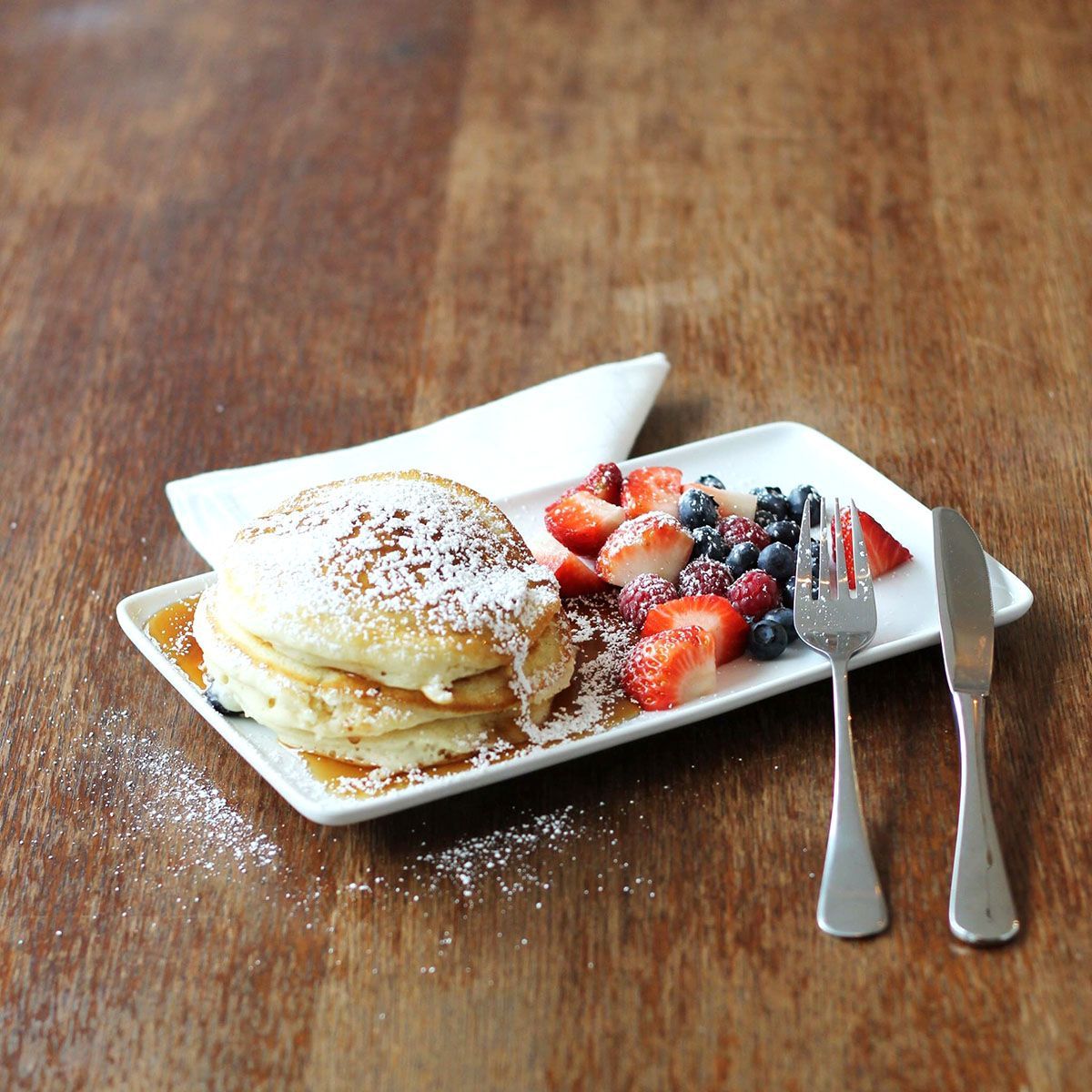 Pannenkoeken opgestapeld op een wit bord met bessen en siroop, bestrooid met poedersuiker, geplaatst op een houten tafel met zilveren bestek.