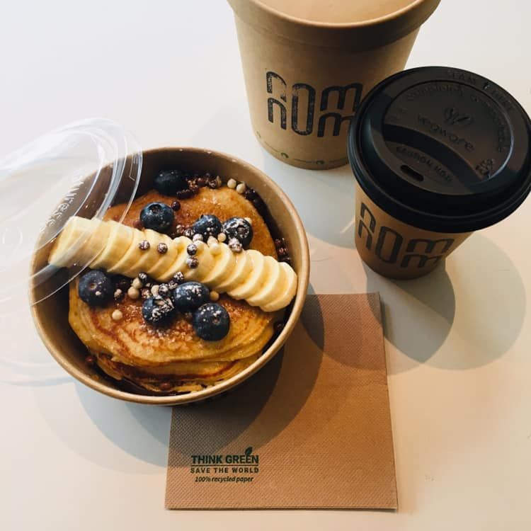 Pannenkoeken met bosbessen en banaan in een kom, twee koffiekopjes en een servet op een witte ondergrond.