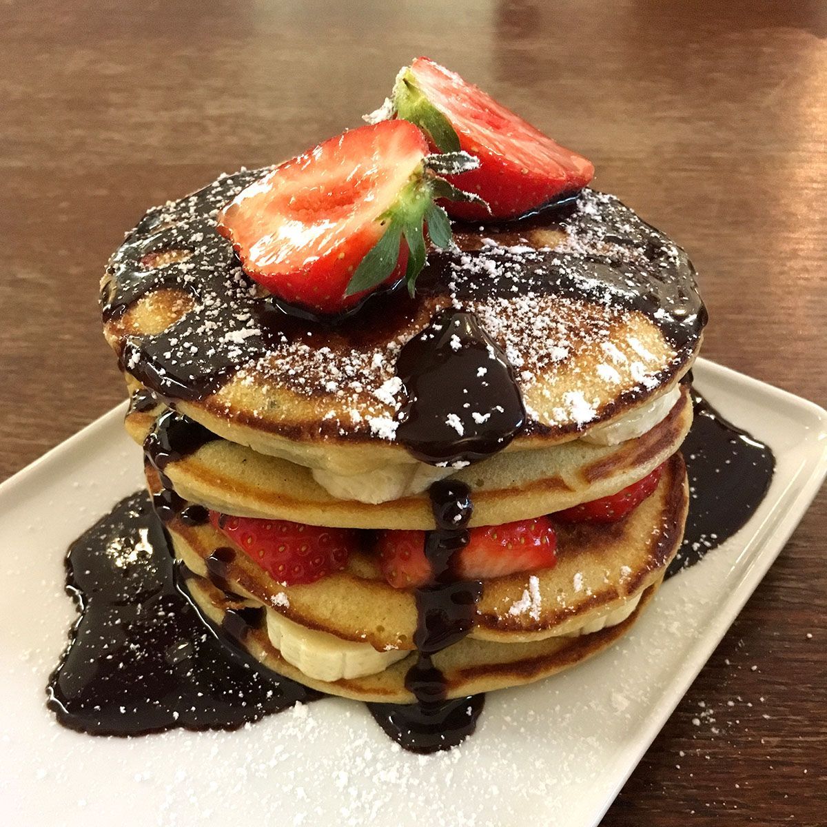Een stapel pannenkoeken met chocoladesaus, aardbeien en bananen, bestrooid met poedersuiker, op een wit bord.