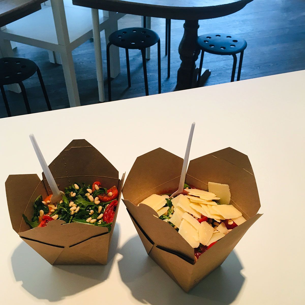 Twee bakjes met salade en een lepel staan op een witte tafel. Op de achtergrond zijn stoelen en een tafel te zien.