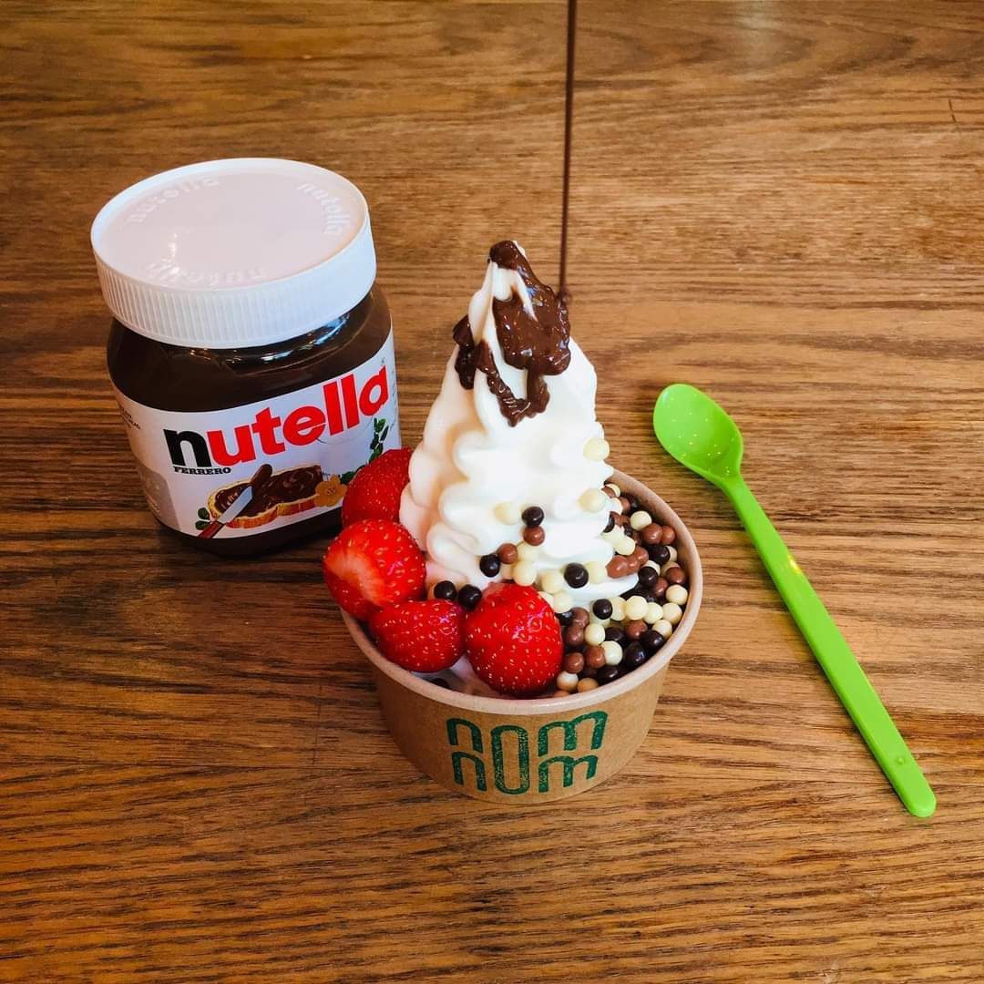 Een bakje bevroren yoghurt met aardbeien, chocoladesprinkels en Nutella, dat wordt besprenkeld met nog meer Nutella; naast een pot Nutella en een groene lepel.