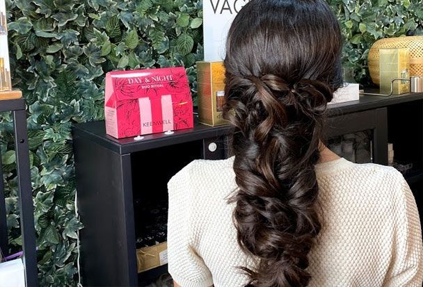 Mujer con cabello oscuro con un peinado trenzado, de pie frente a un mostrador con una caja de productos de belleza.