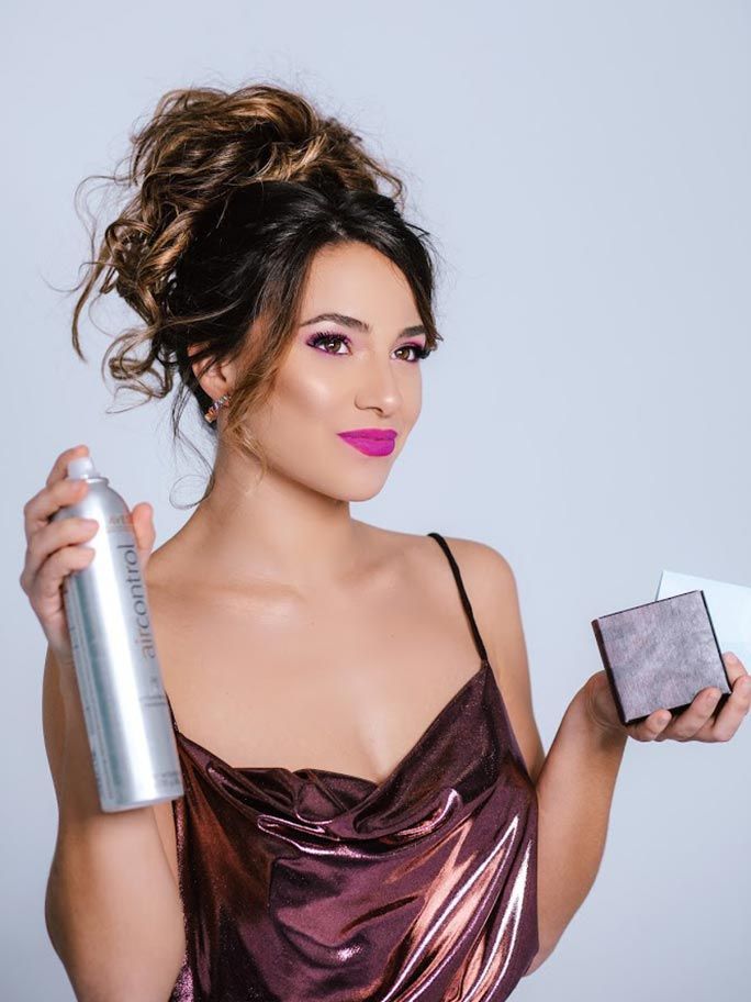 Mujer con un vestido brillante sostiene laca para el cabello y maquillaje compacto, mirando a la cámara.